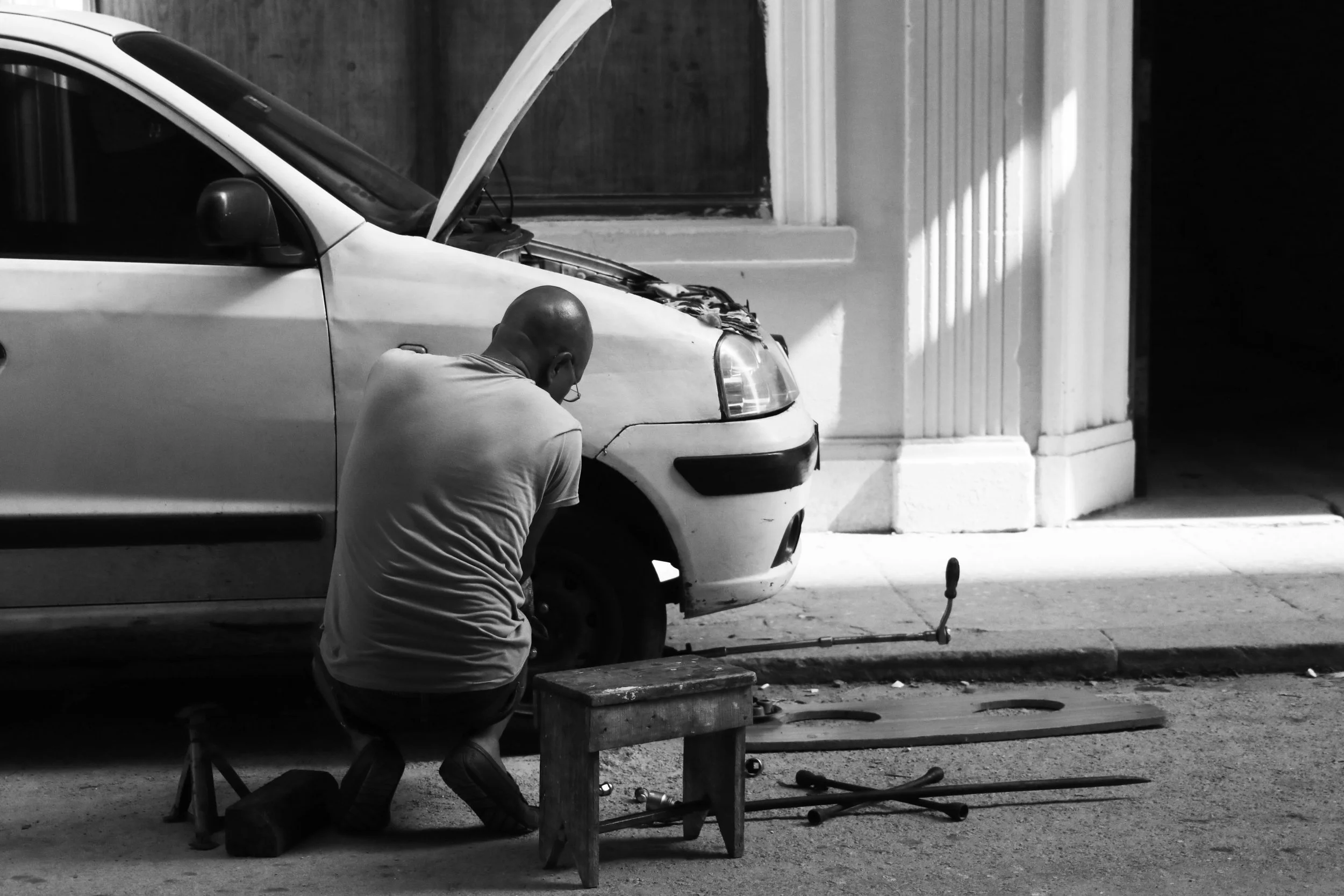 A man working on a car with the hood open on an urban street, with tools and a small wooden stool nearby.