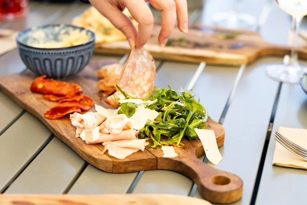 Person placing slices of salami on a wooden serving board with greens, sliced turkey, and crispy bacon, with a bowl of potato salad and wine glasses in the background.