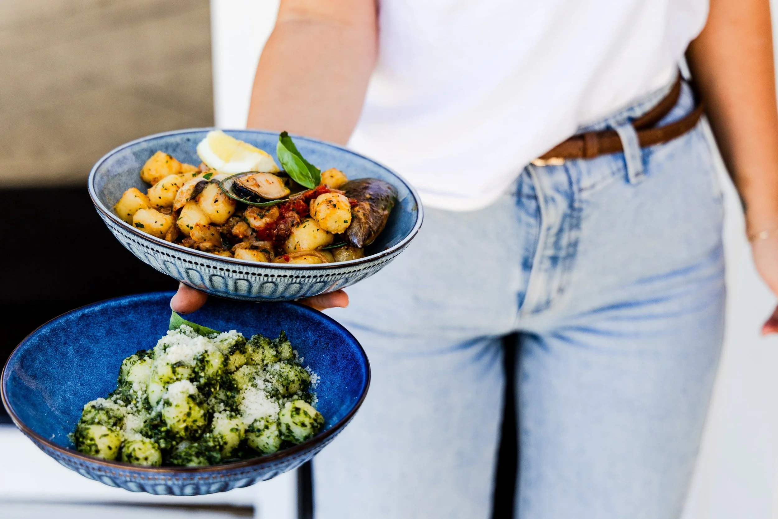 Person holding a blue bowl of gnocchi with herbs and a lemon wedge, wearing a white shirt and blue jeans.