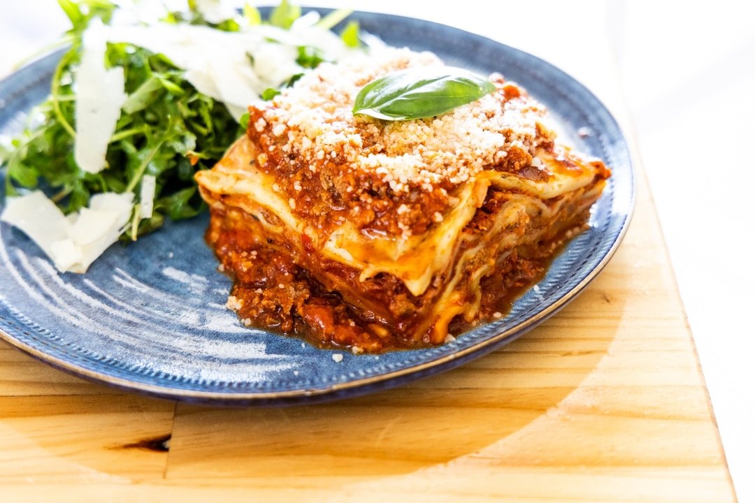 A serving of lasagna on a blue plate with shredded cheese and a basil leaf on top, accompanied by a side of salad.