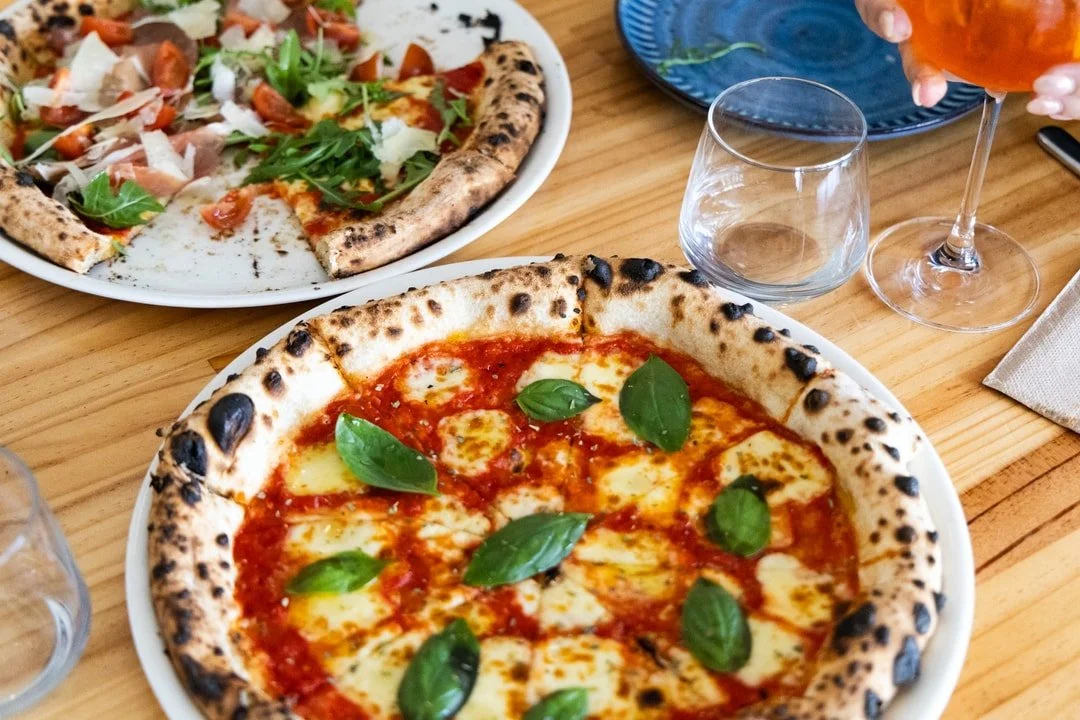 Two pizzas on a wooden table, one with fresh mozzarella, tomato sauce, basil leaves, and the other with arugula, cherry tomatoes, and shaved cheese, alongside empty glasses and a glass of rosé wine.