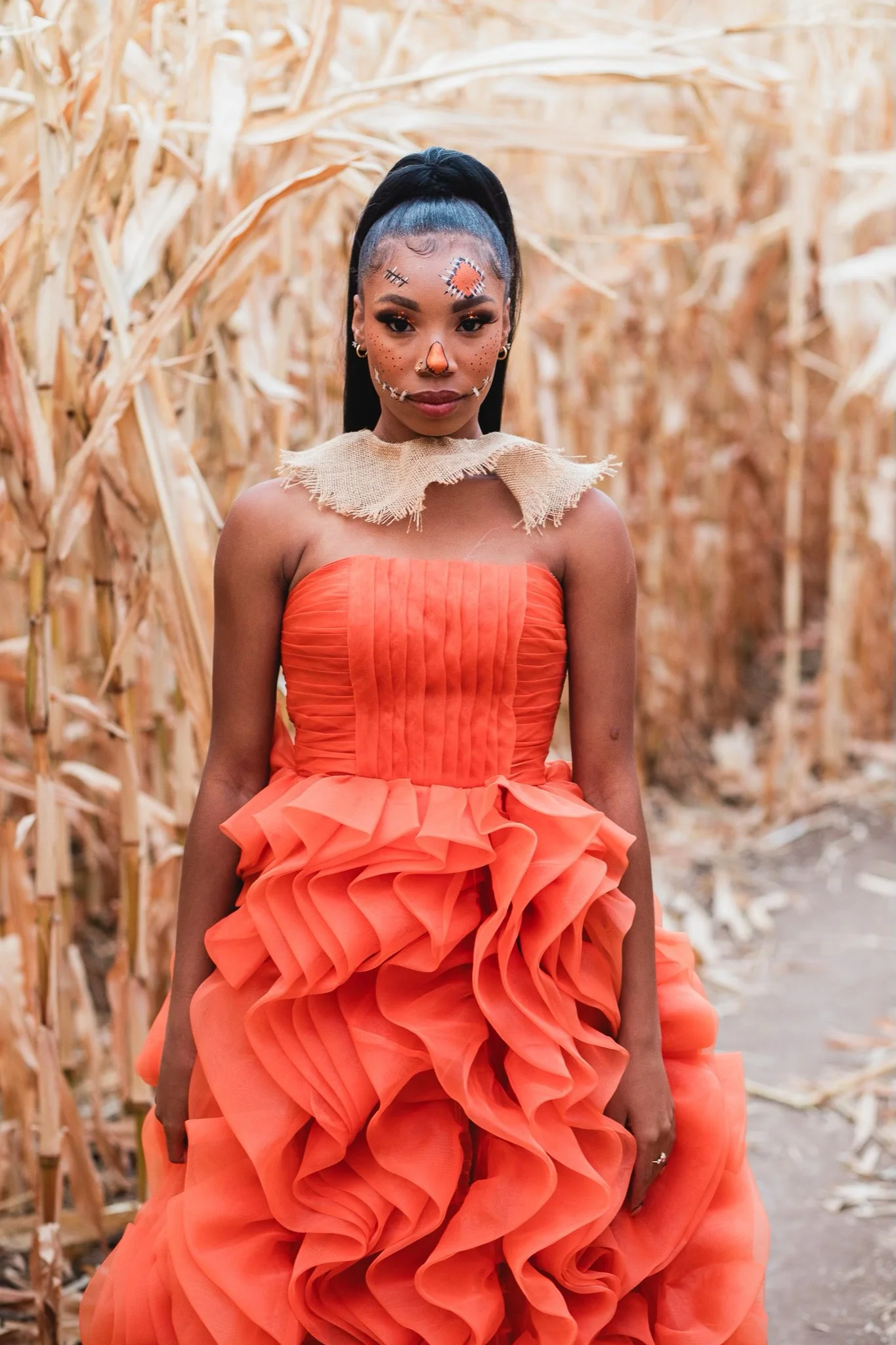 A woman in an orange dress with ruffled layers standing in a cornfield with dried corn stalks behind her.