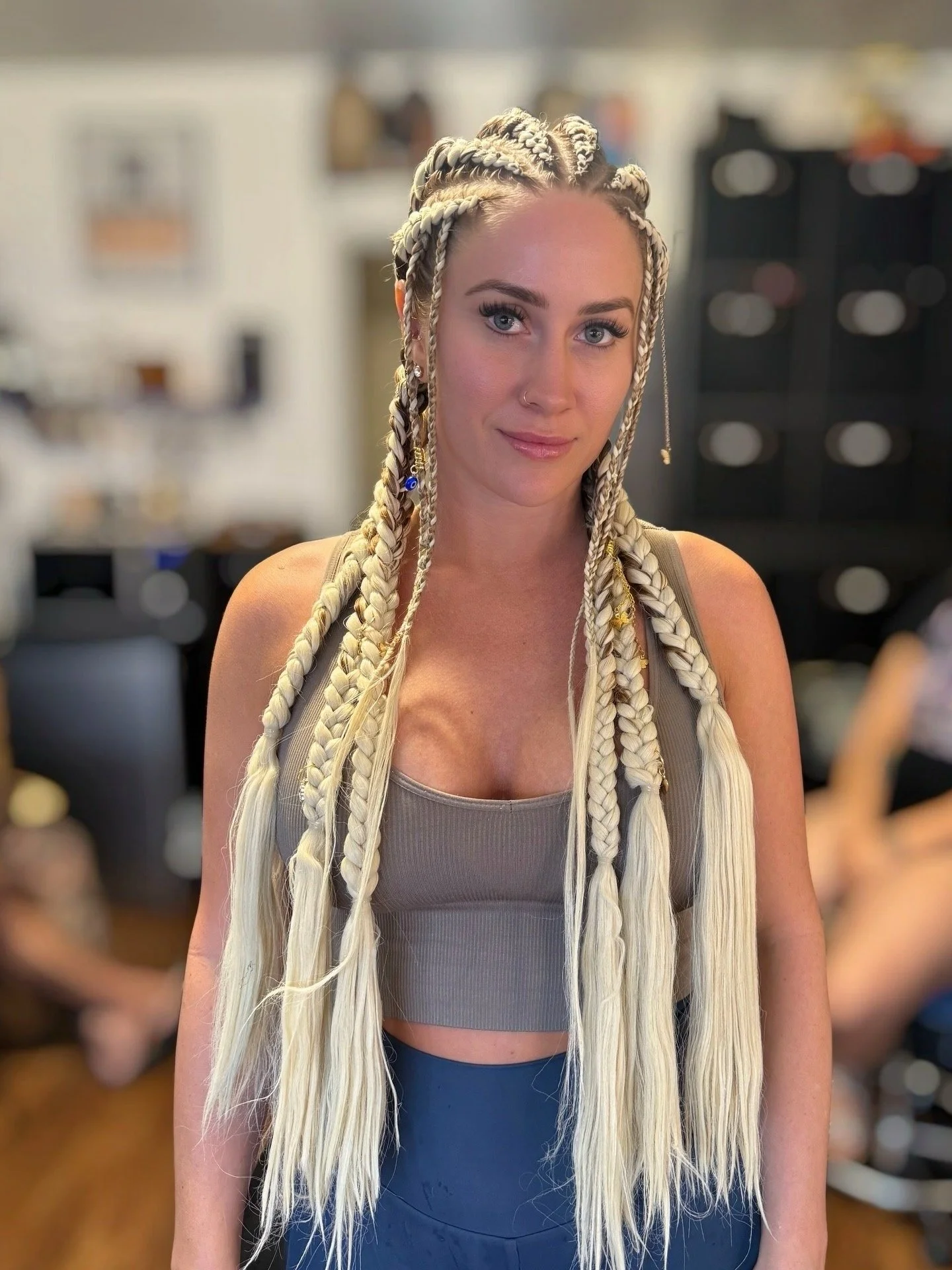 A woman with blonde braids with beads, wearing a gray tank top and blue leggings, standing indoors with a blurred background.