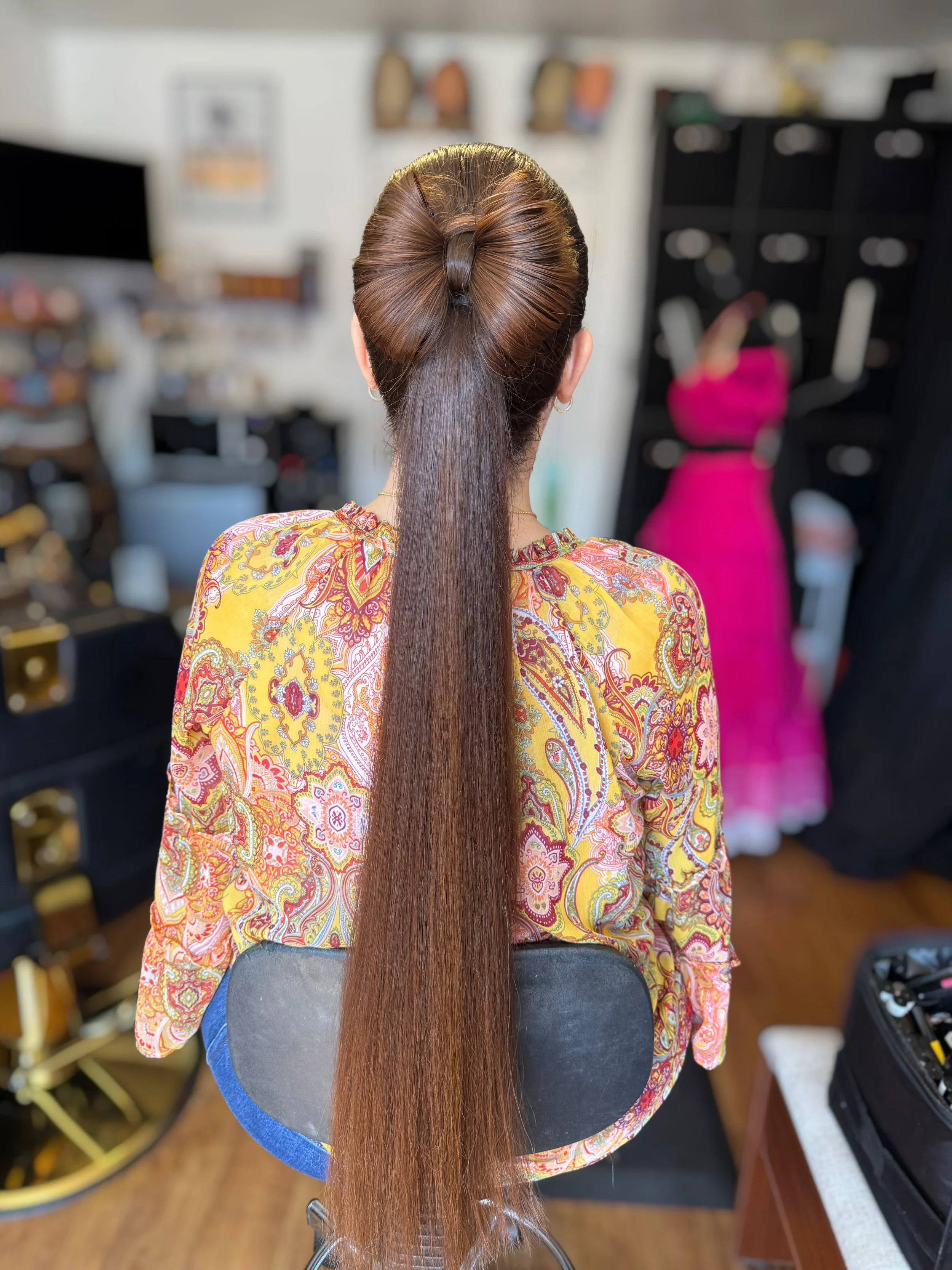 Back view of a woman with long, straight, brown hair sitting in a salon chair with a half-up, half-down hairstyle, wearing a floral-patterned shirt.