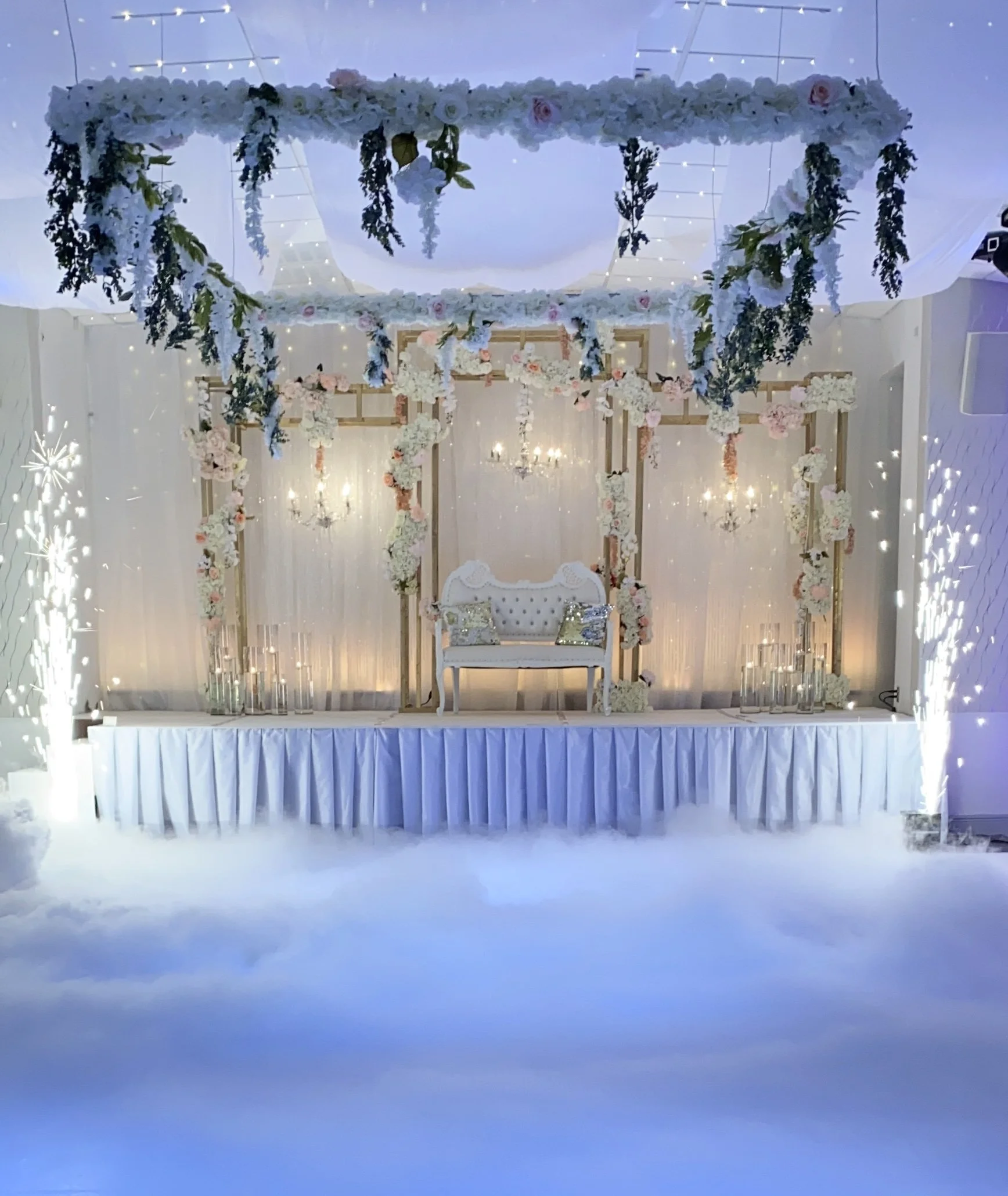 Decorative stage with white loveseat, floral arrangements, and chandeliers, set against a white backdrop with drapes and soft lighting.