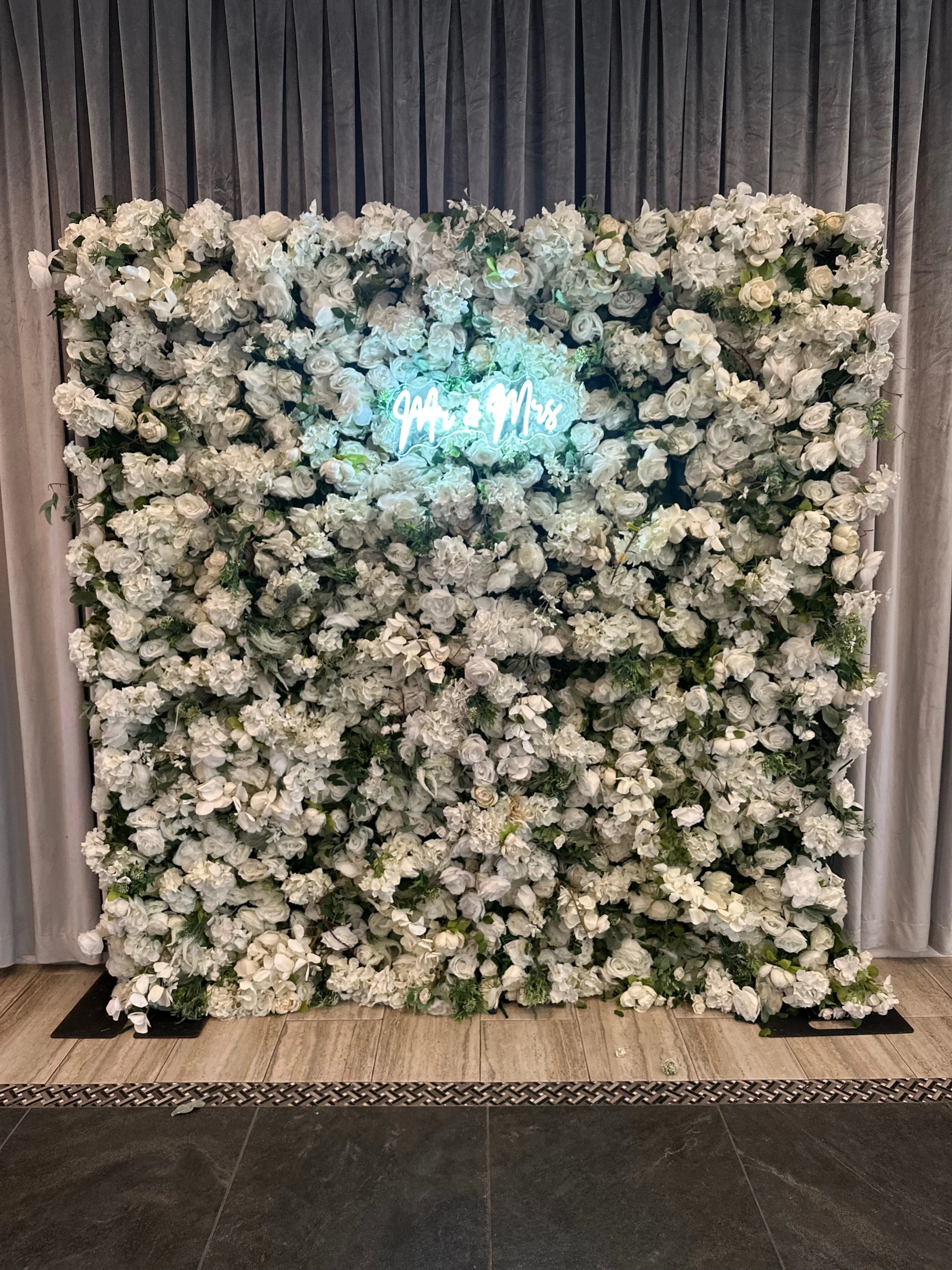 A floral wedding backdrop with white roses and green foliage, featuring a neon sign that reads 'Mr. & Mrs.' in the center, positioned in front of gray curtains.
