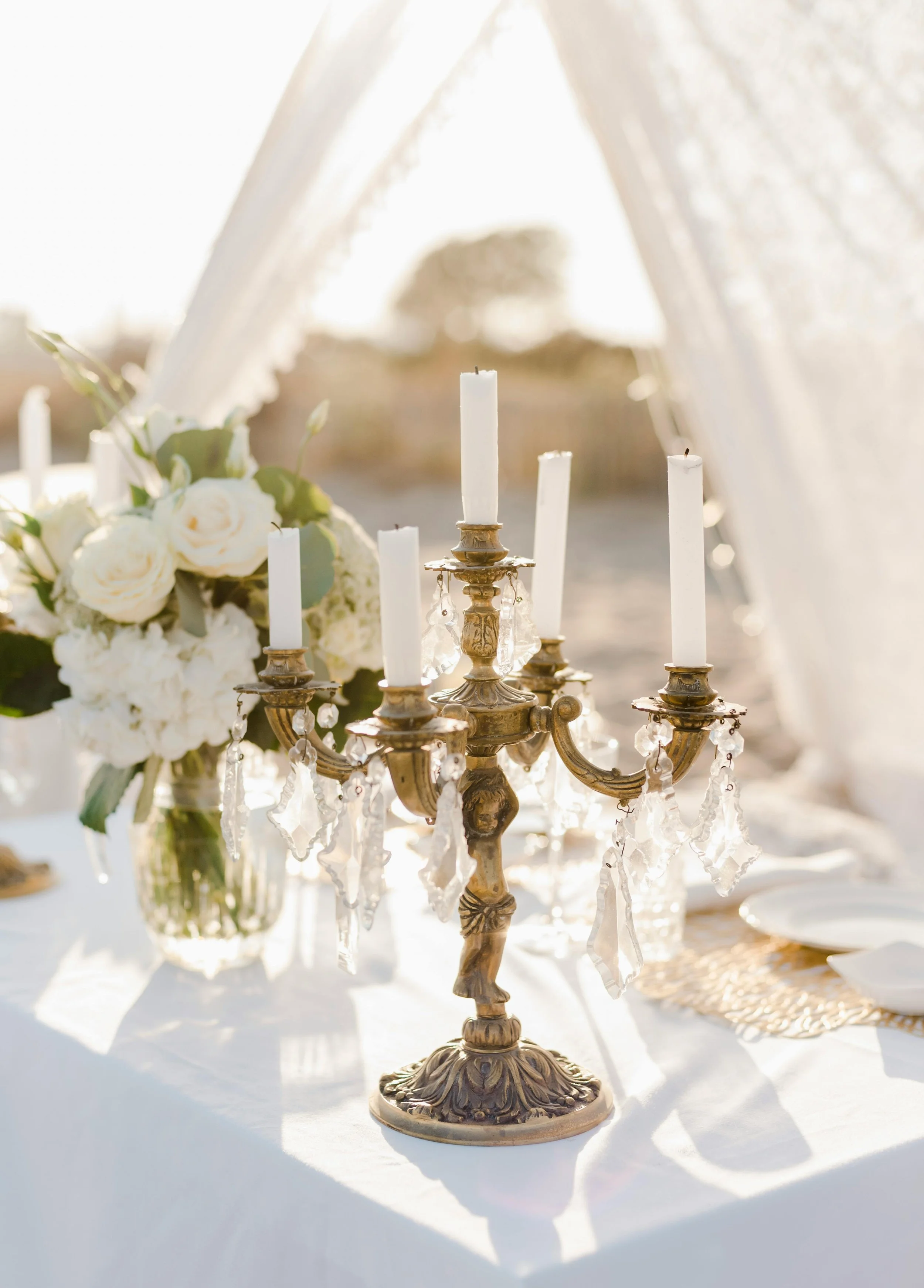 A vintage brass candelabra with five white candles, decorated with hanging crystals, placed on a table with a white tablecloth and a floral arrangement in a glass vase, outdoors with sunlight.