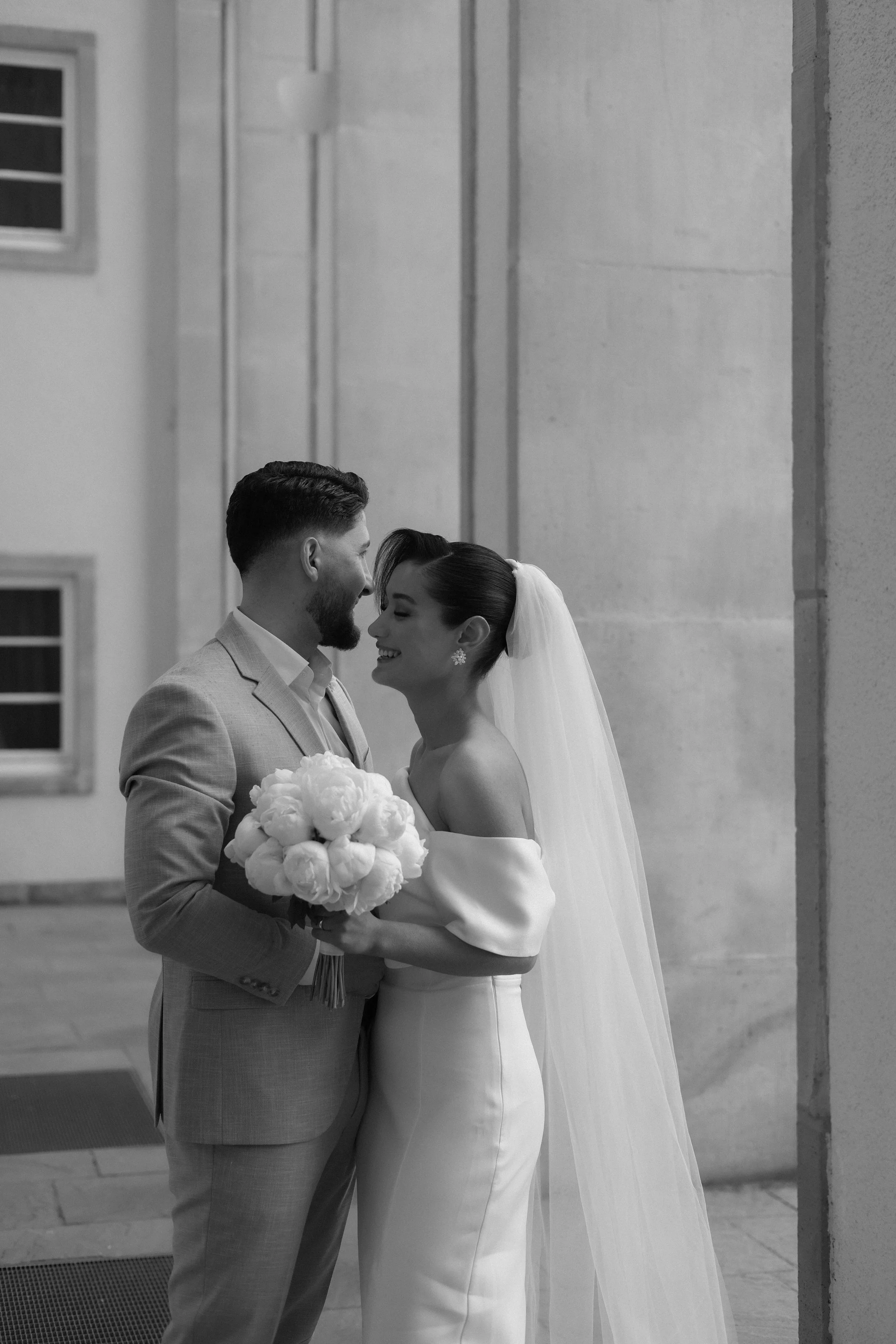 A black and white photo of a bride and groom smiling and holding a bouquet, standing close together in a building with concrete walls and windows.