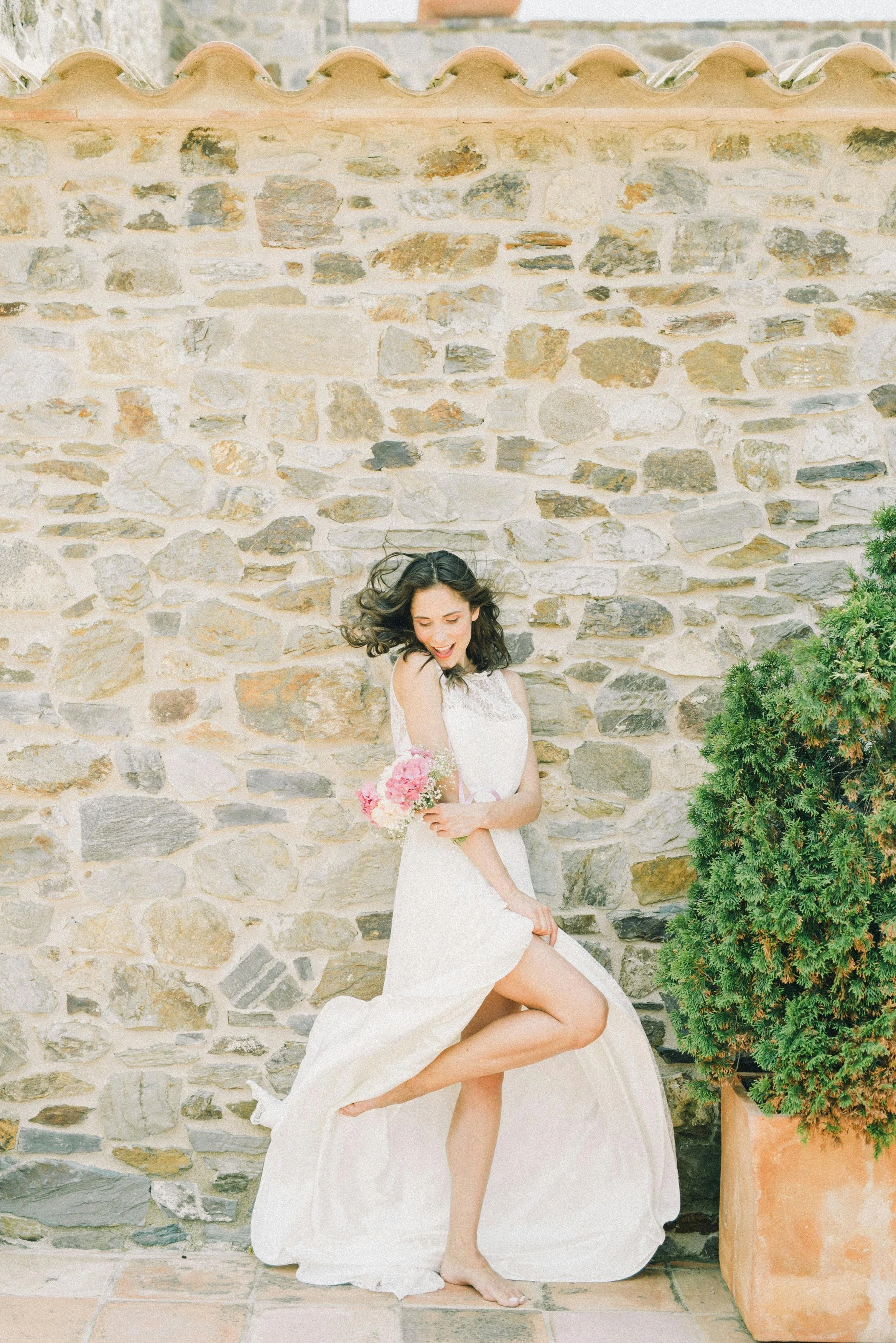 Woman in a white dress with a pink flower bouquet dancing and smiling against a stone wall background.