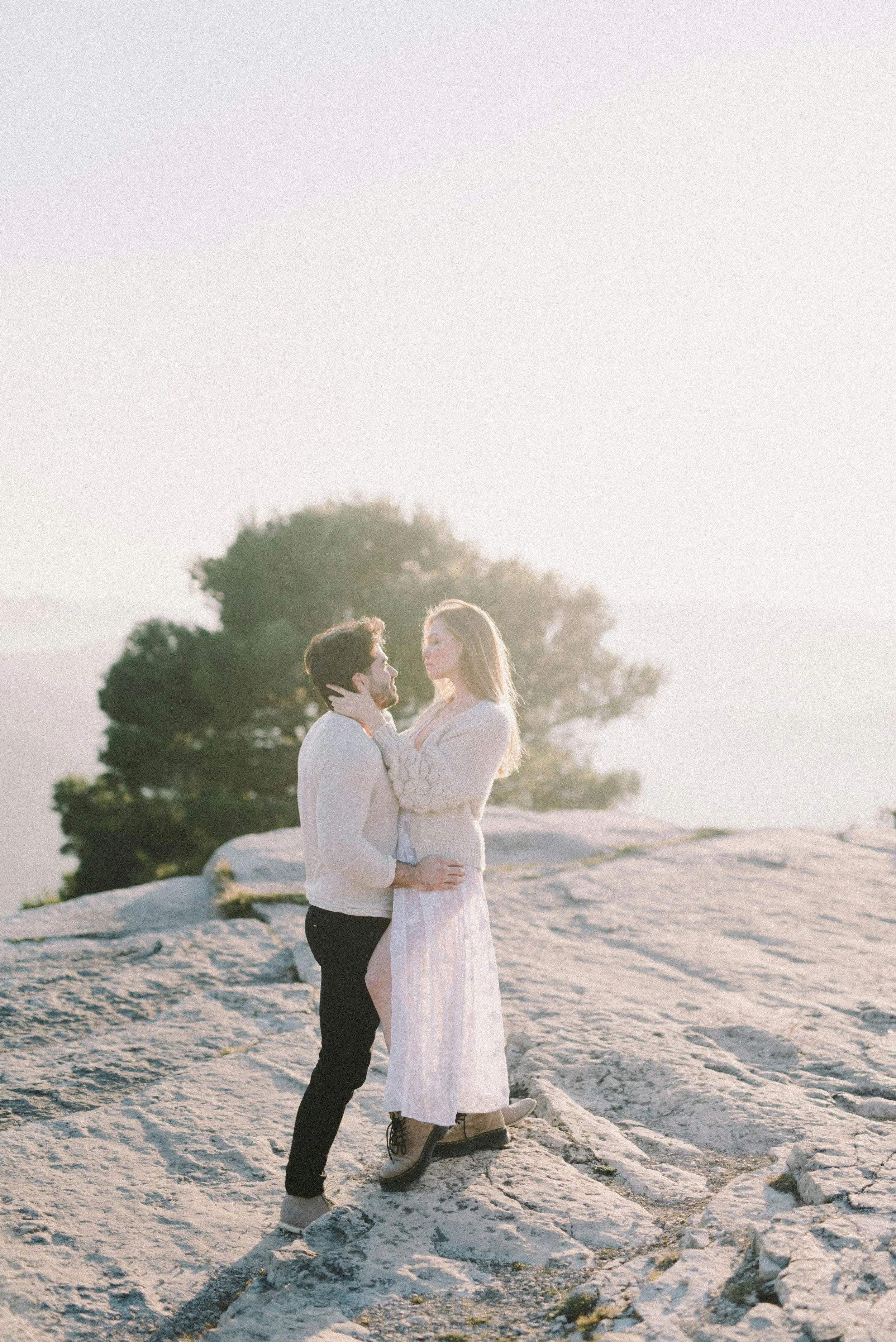 A couple standing on rocks outdoors, facing each other tenderly, with sunlight in the background.