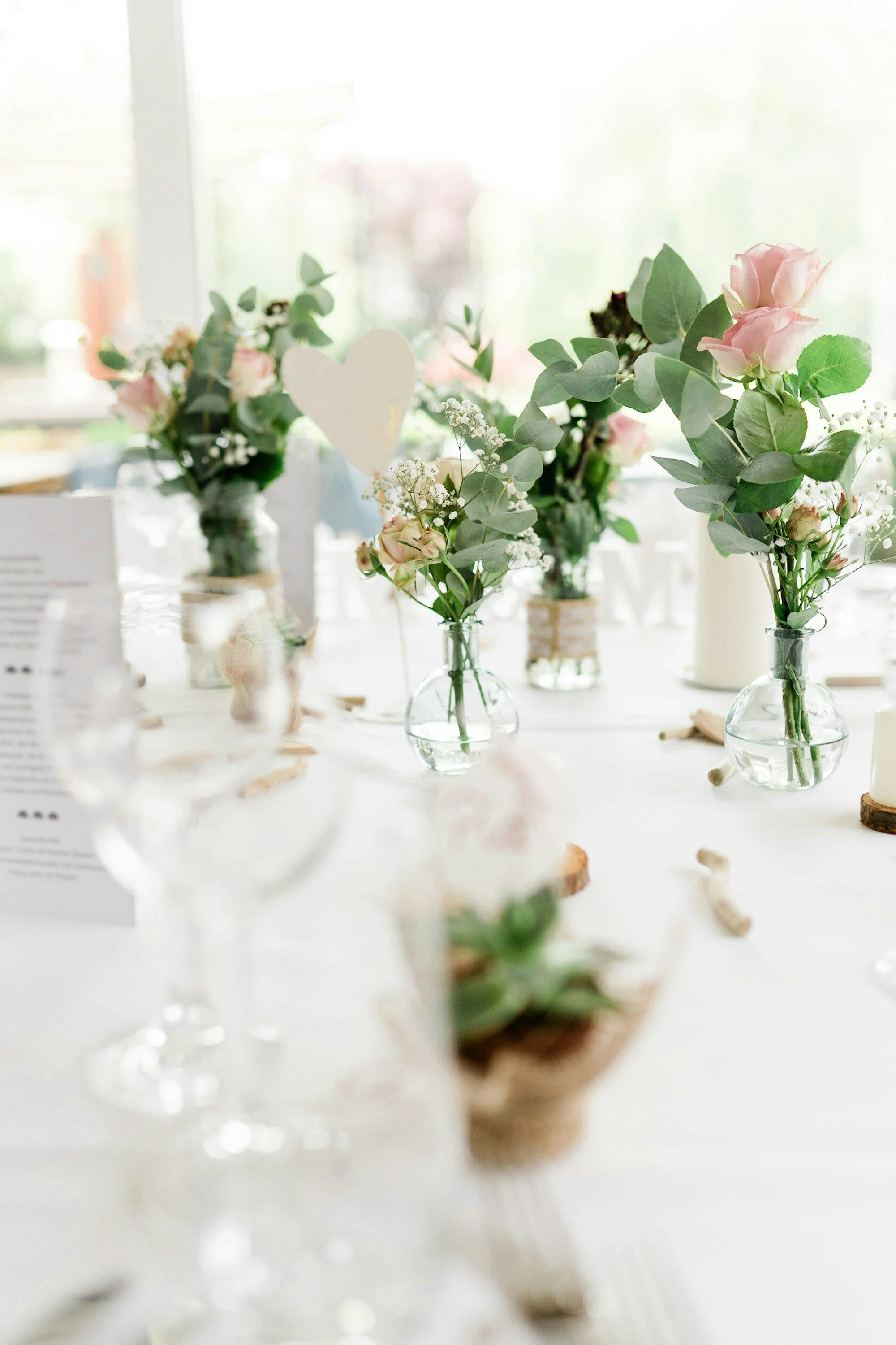 Elegant table setting with various small glass vases filled with light pink roses, greenery, and white flowers, placed on a white table with candles and decorative elements.