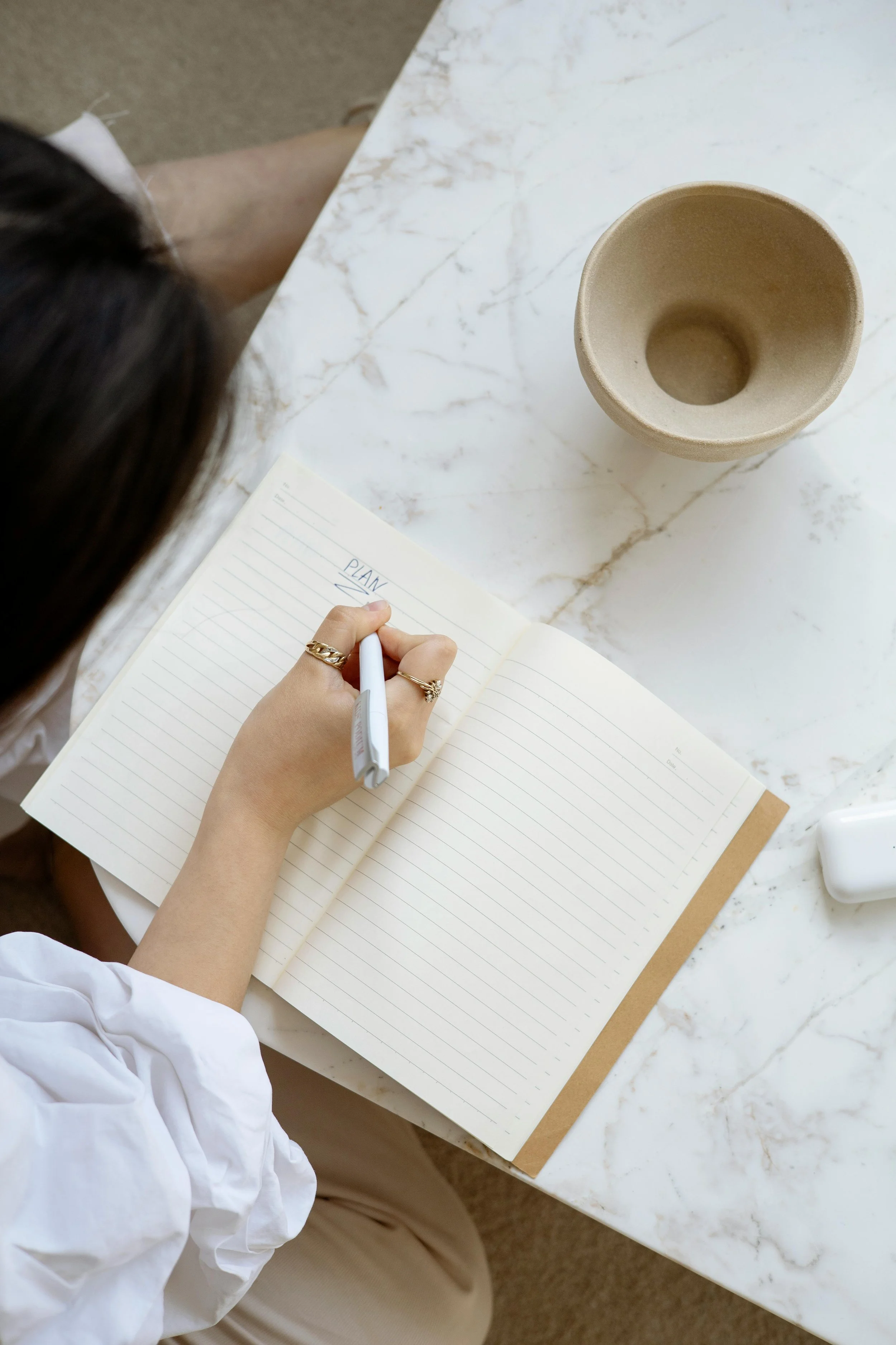 A person is writing in a notebook with the word 'PLANNING' at the top, sitting at a white marble table with a round beige cup and a white object nearby.