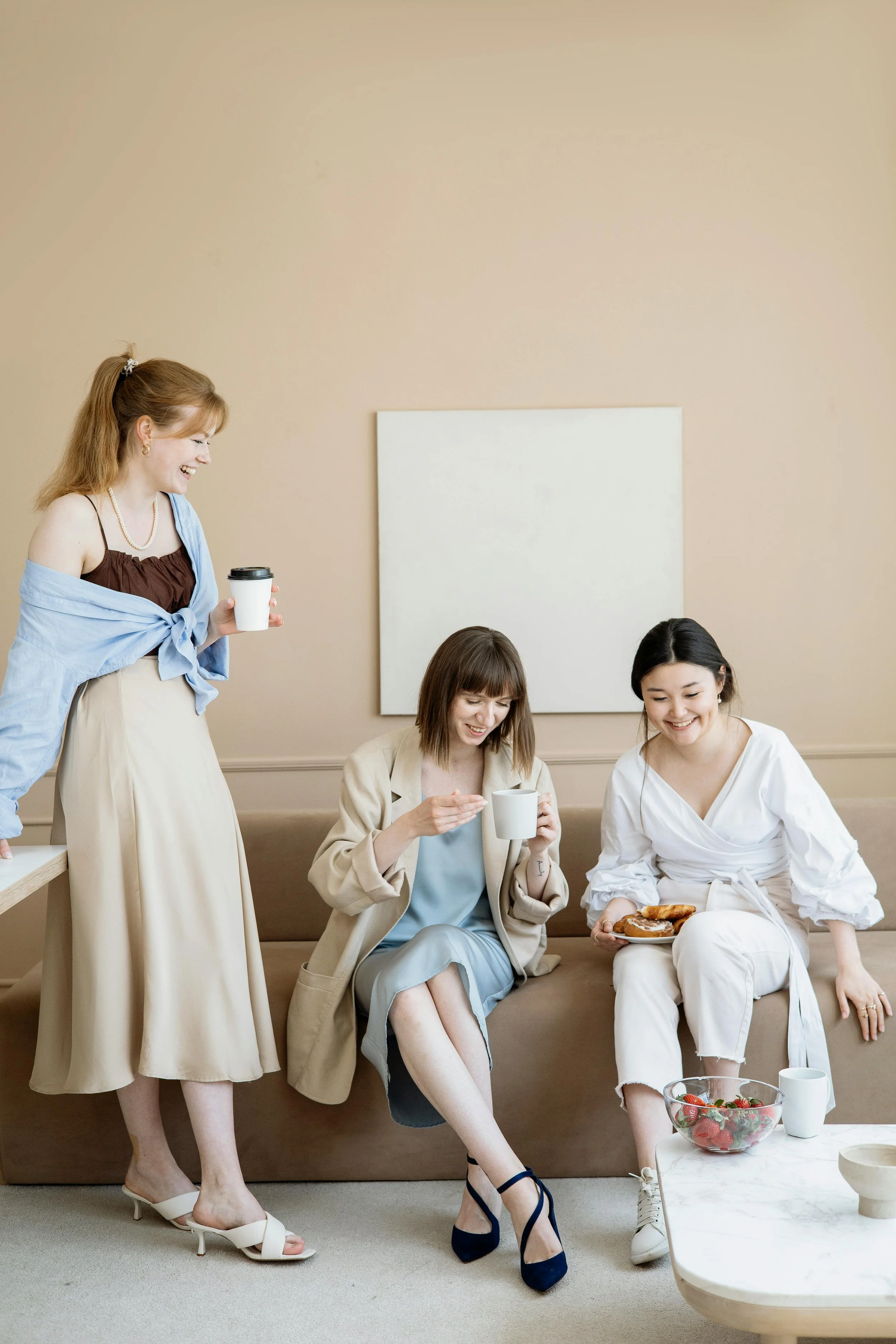 Three women laugh and share coffee and snacks in a cozy living room with beige walls and minimalist decor.