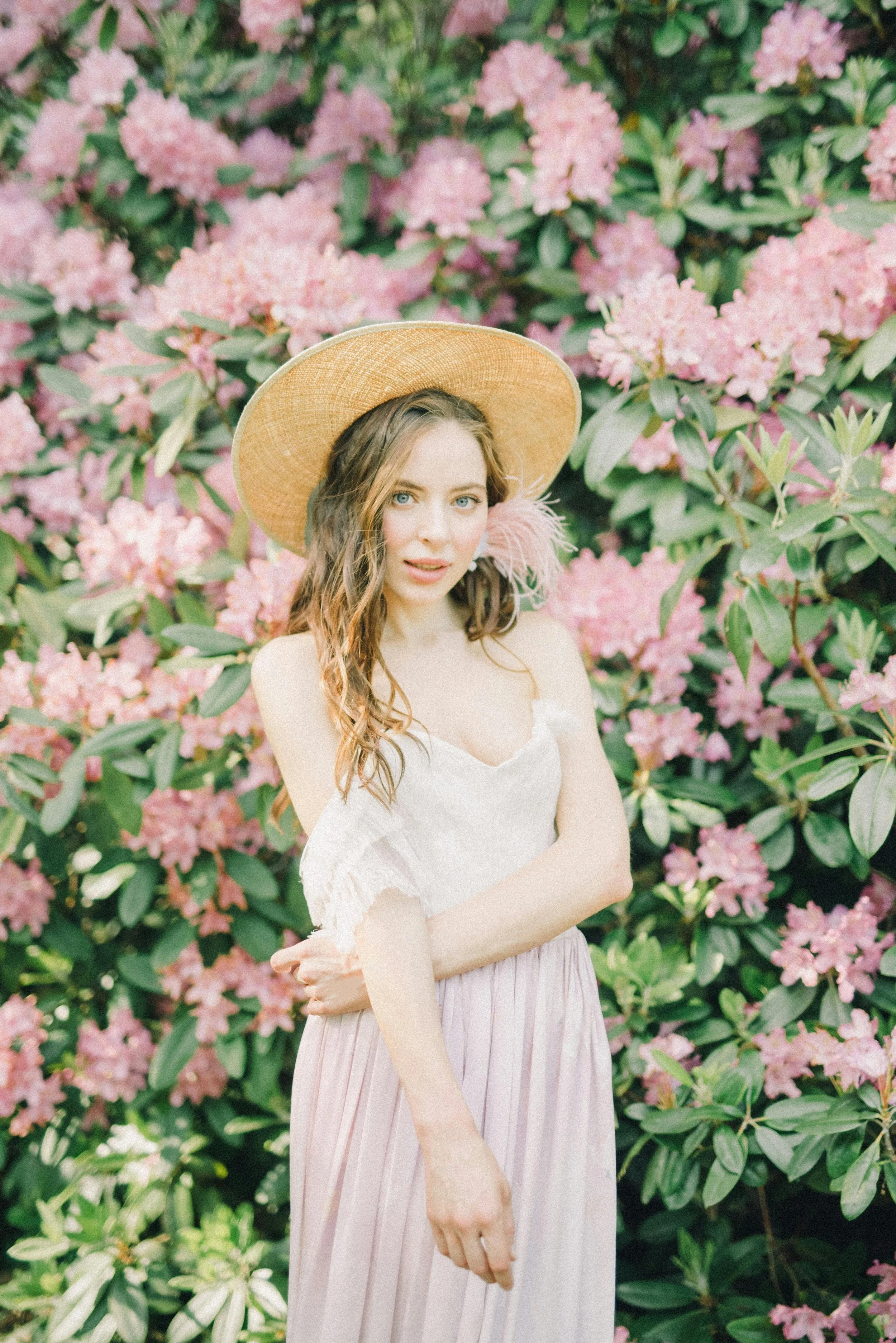 A young woman with wavy brown hair and blue eyes standing in front of pink flowering bushes, wearing a wide-brimmed straw hat with a pink feather accent, a white off-shoulder top, and a light pink pleated skirt.
