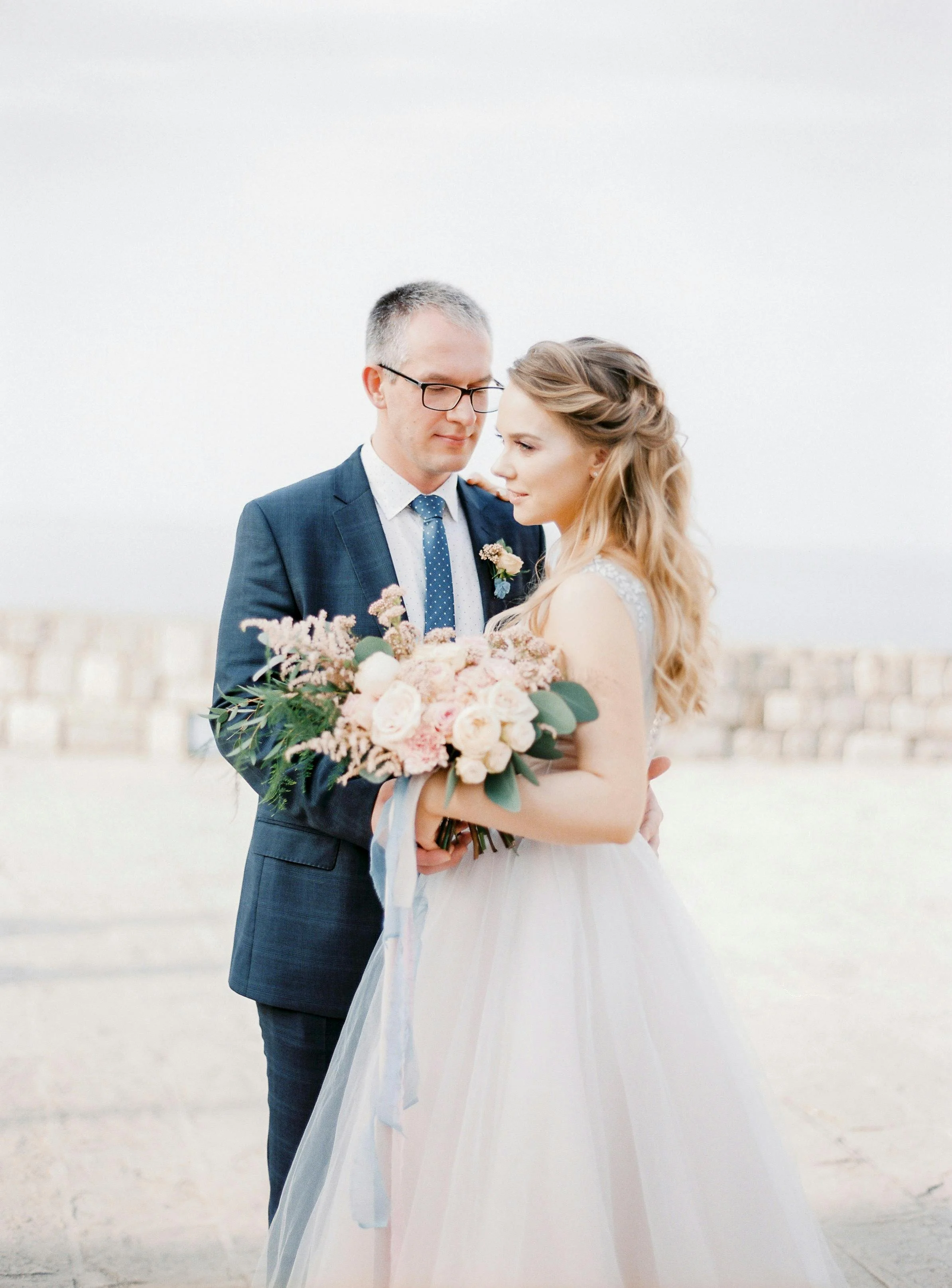 A bride holding a bouquet of pink and white flowers, standing next to a groom wearing a navy suit, during a wedding photoshoot outdoors.