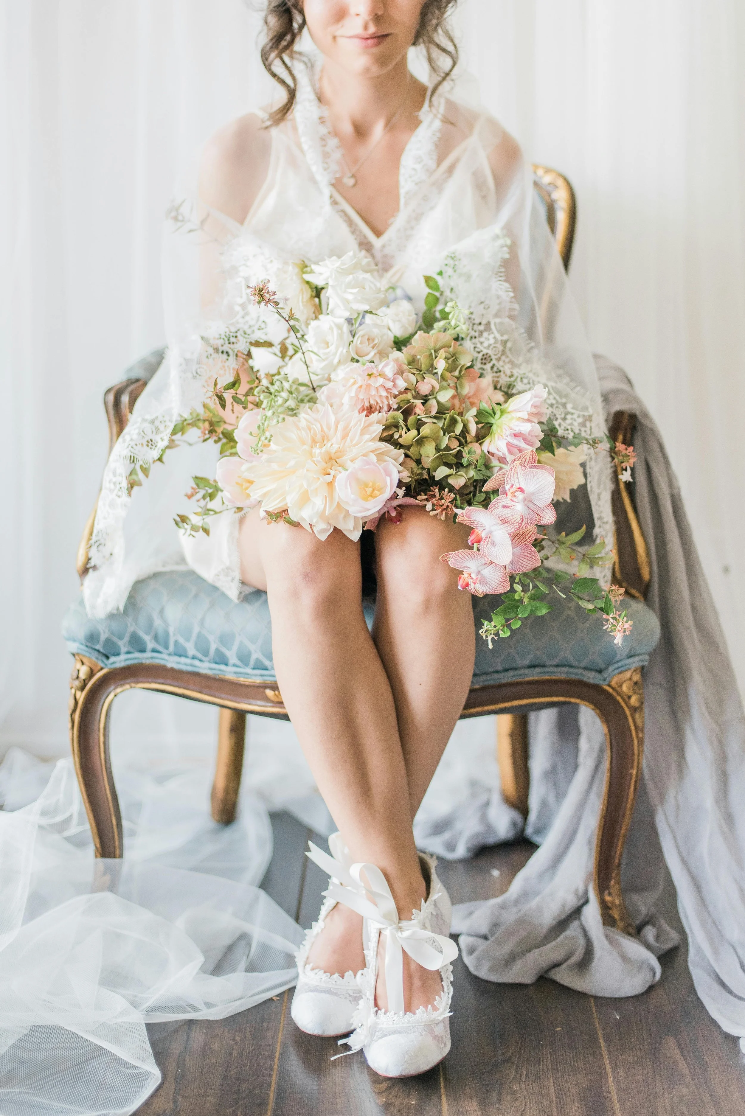 A woman wearing white lace shoes with ribbon ties is seated with crossed legs, holding a large bouquet of pink and white flowers, dressed in a light, delicate, lace-looking outfit, sitting on a vintage, blue upholstered chair with gold accents, surrounded by soft, sheer fabric and a gray cloth draped on the side.