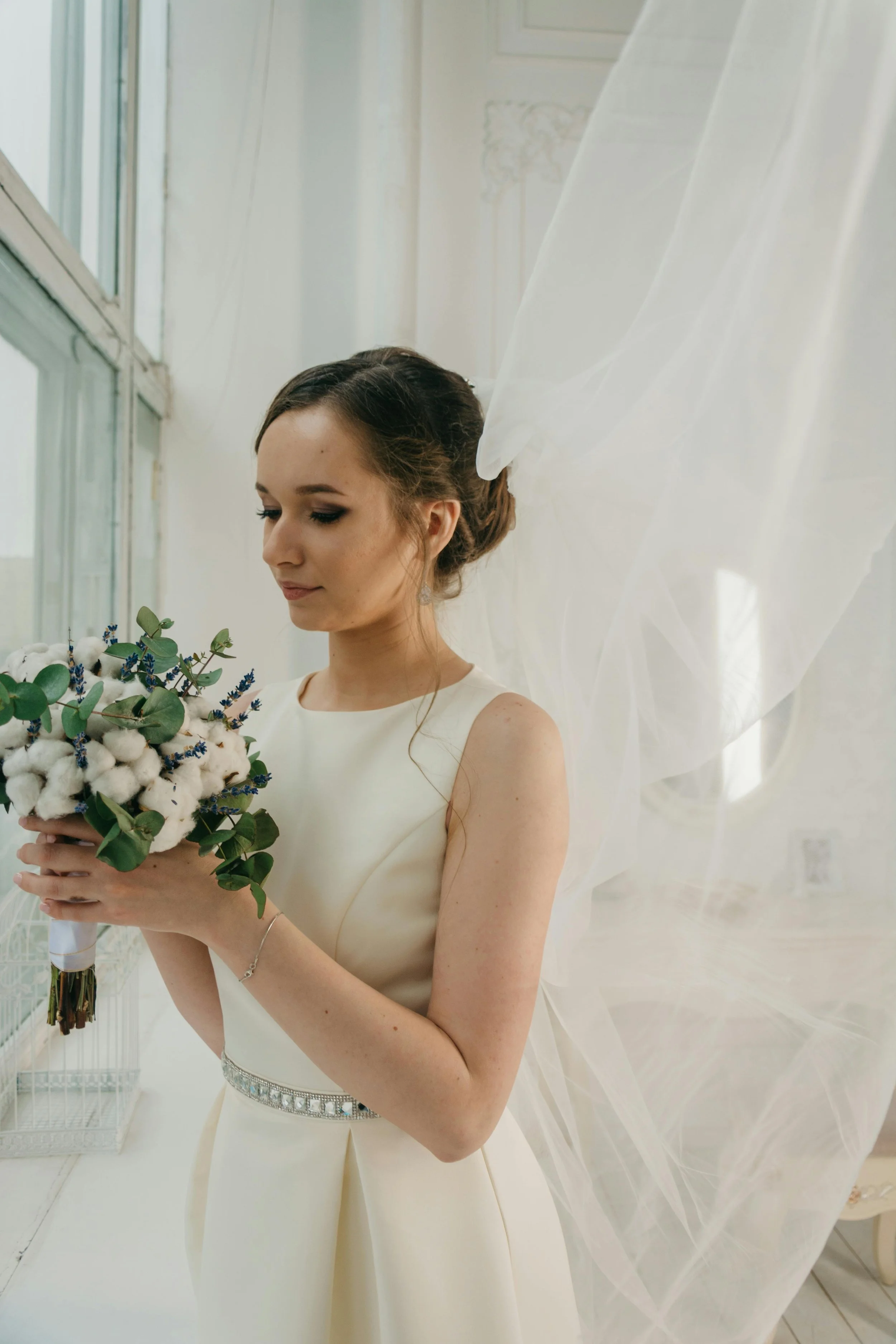 A bride in a cream-colored dress holding a bouquet of cotton, eucalyptus, and lavender, standing beside a window with sheer white curtains.
