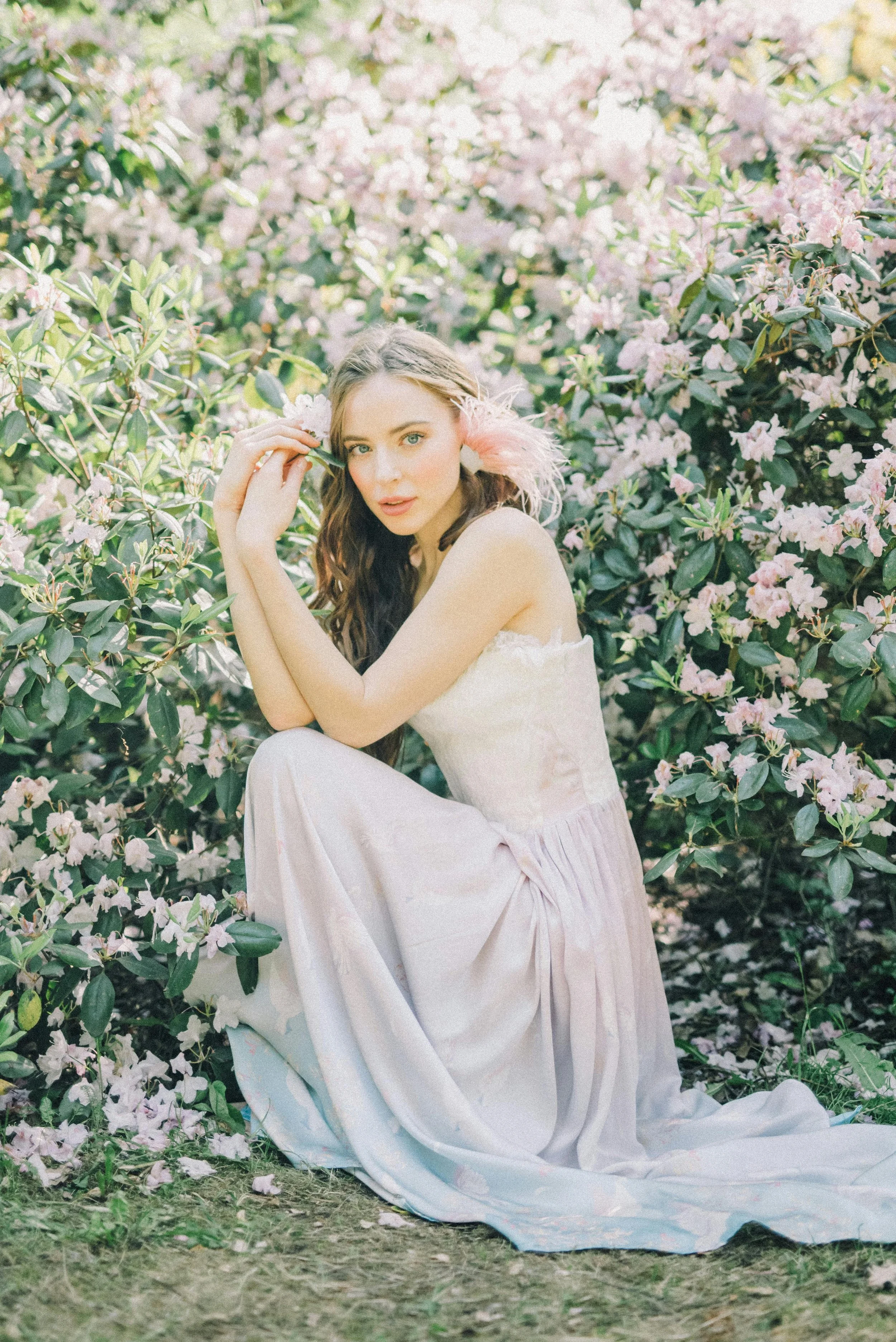 A young woman with long brown hair, sitting among pink blooming azalea bushes, wearing a strapless white dress with a pink feather accessory in her hair, looking at the camera.
