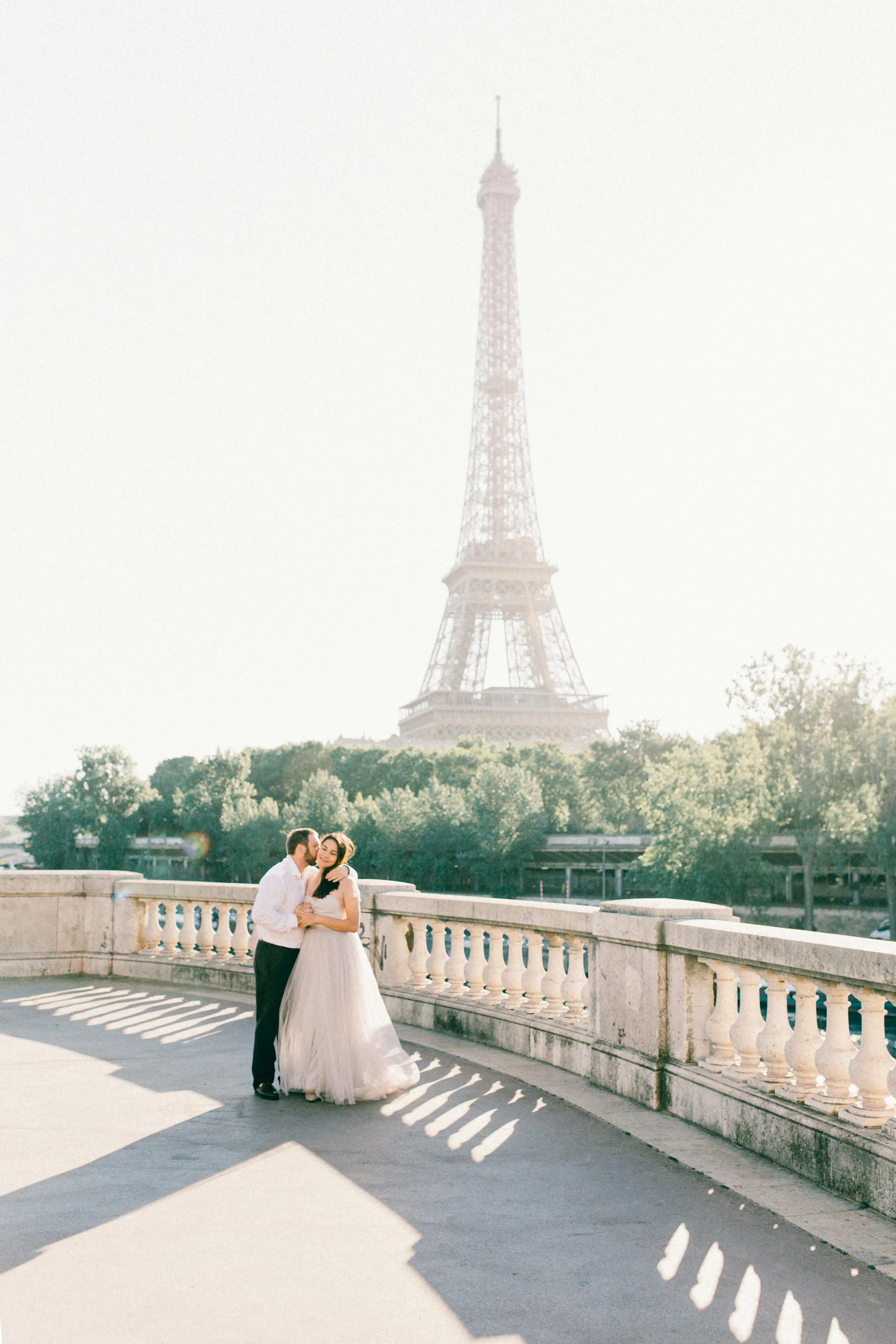 A bride and groom embrace on a bridge with the Eiffel Tower in the background on a sunny day.