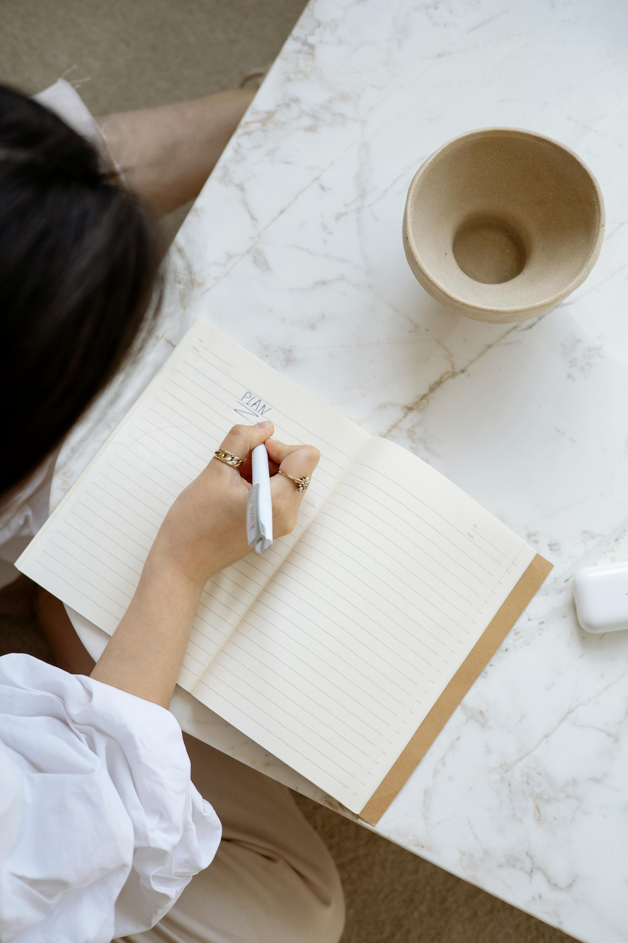 A person writing in a notebook with a white pen on a marble table. The notebook has the word 'PLANS' written at the top of the page. Next to the notebook is a beige empty cup and a white object, possibly a charger or container.
