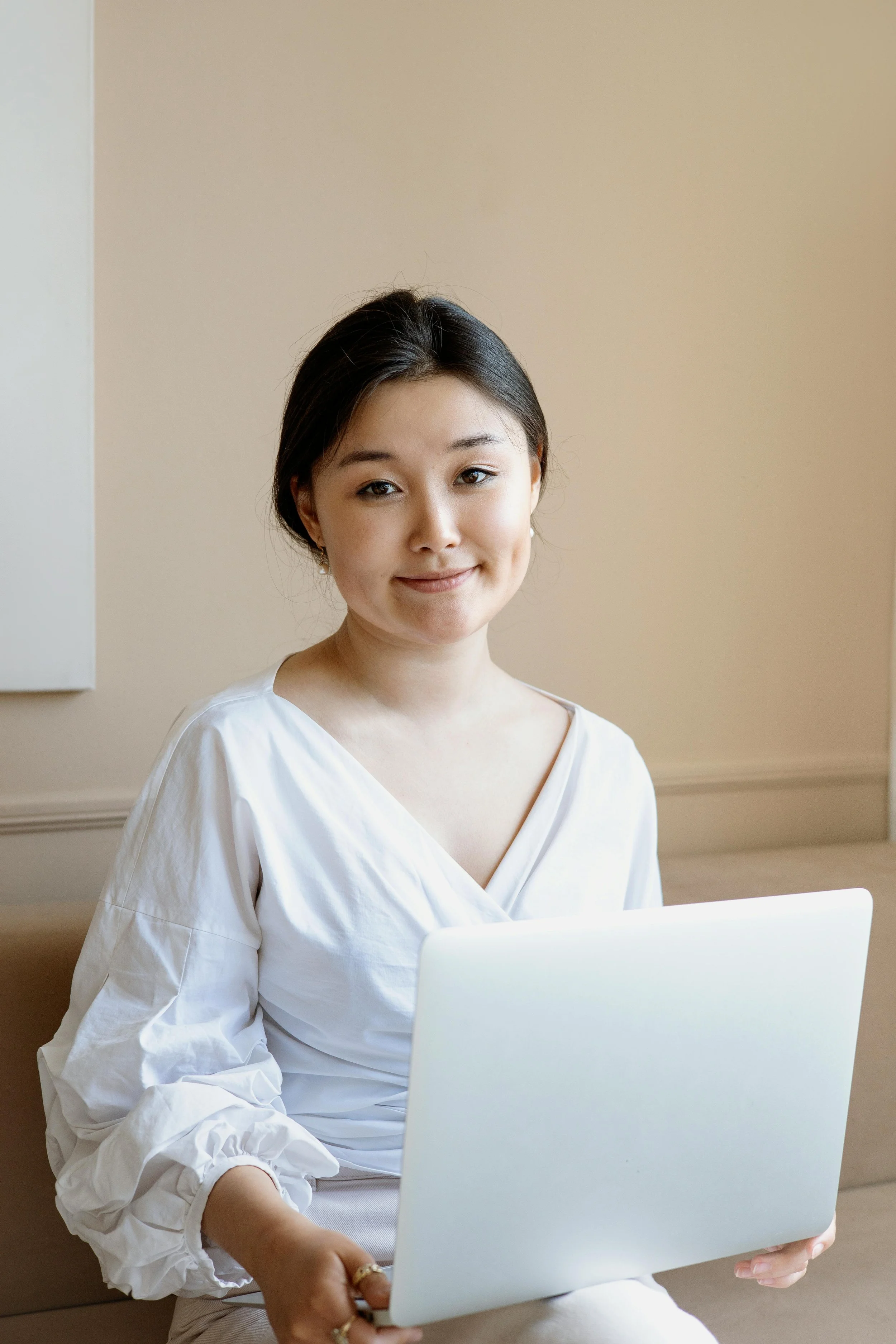 A young Asian woman sitting on a bench, holding a laptop, and smiling at the camera.