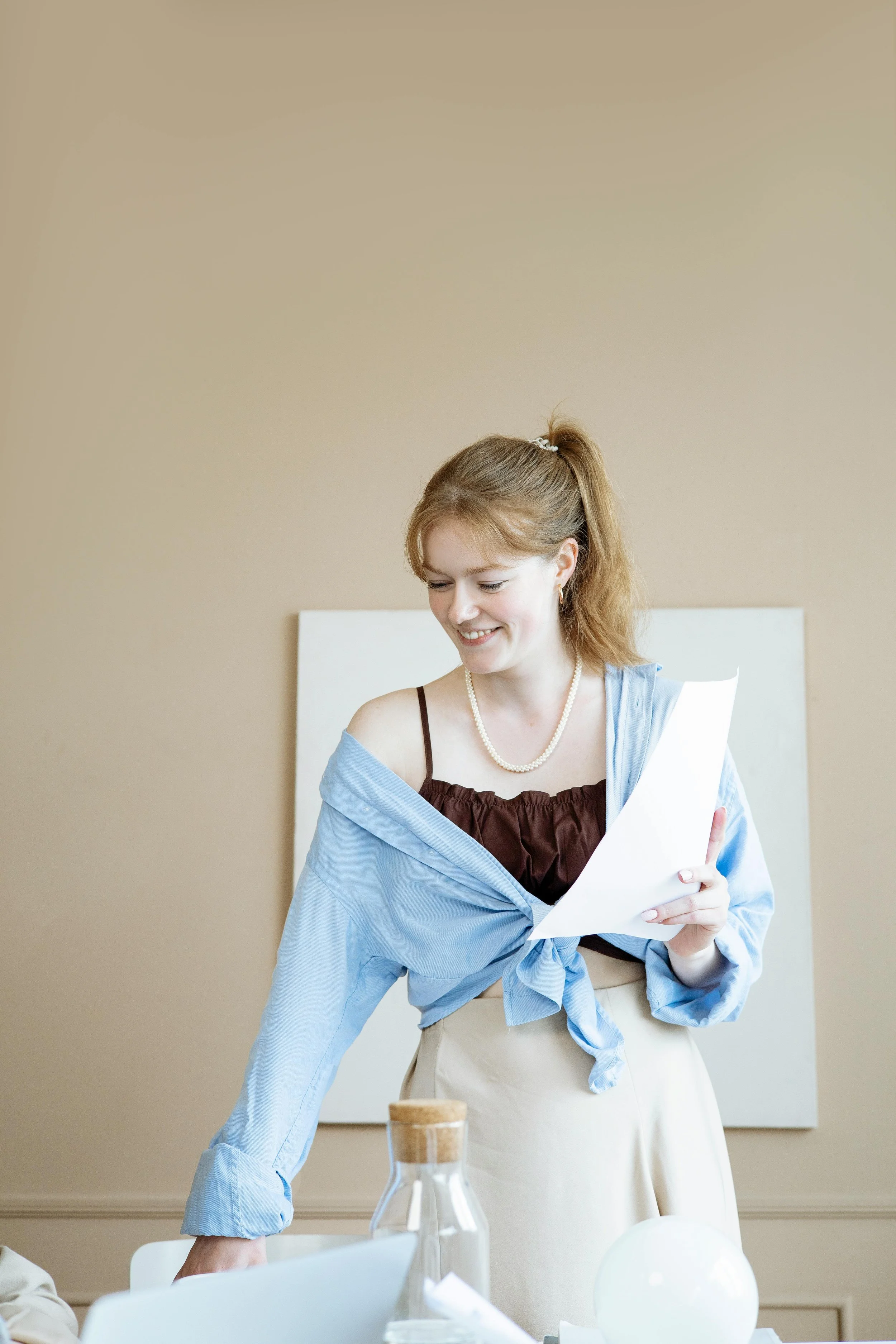 A woman with red hair, wearing a pearl necklace, a brown top, and a blue shirt tied around her waist, is smiling and looking down at a paper she is holding.