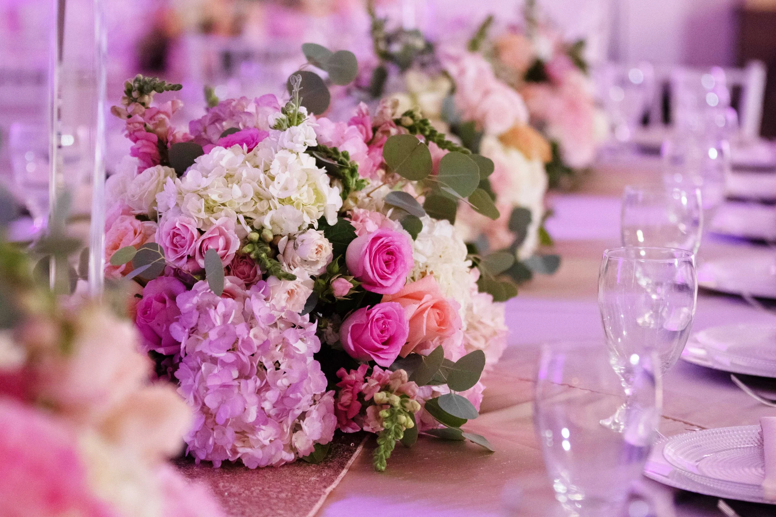 Elegant wedding table centerpiece with pink, white, and peach roses, hydrangeas, and eucalyptus leaves, set on a decorated table with wine glasses and plates.