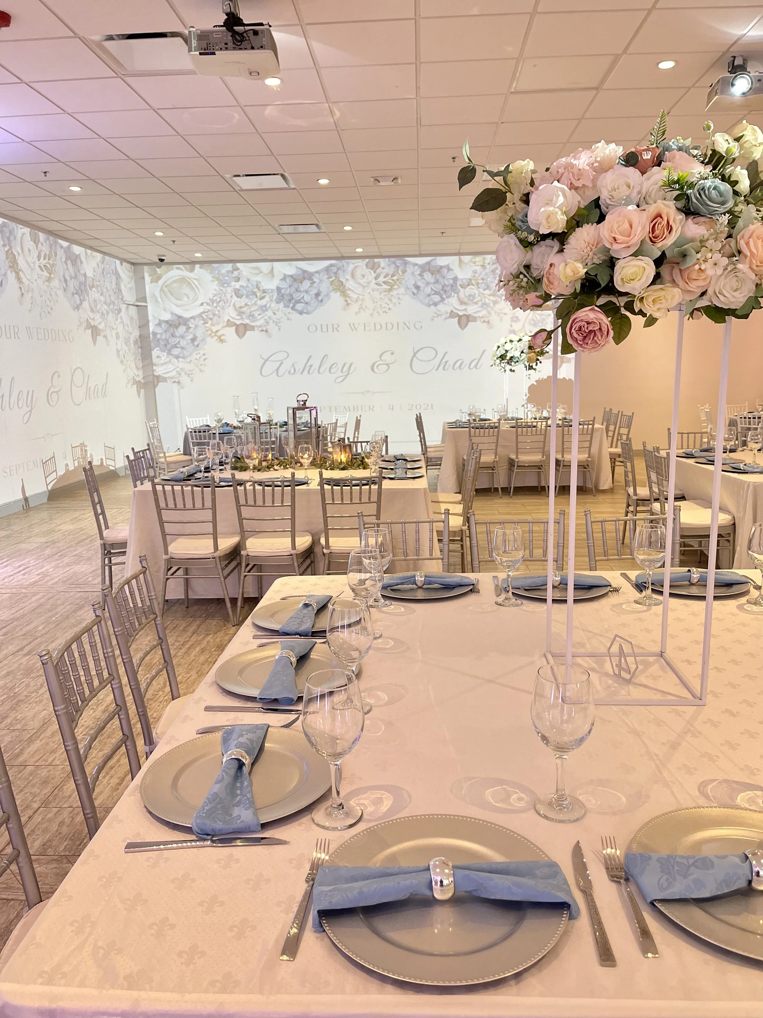 Elegant wedding reception with round and rectangular tables decorated with pastel blue napkins, glassware, and floral centerpieces featuring pink and white roses, in a decorated banquet hall with a lit backdrop displaying the names Ashley and Chad.