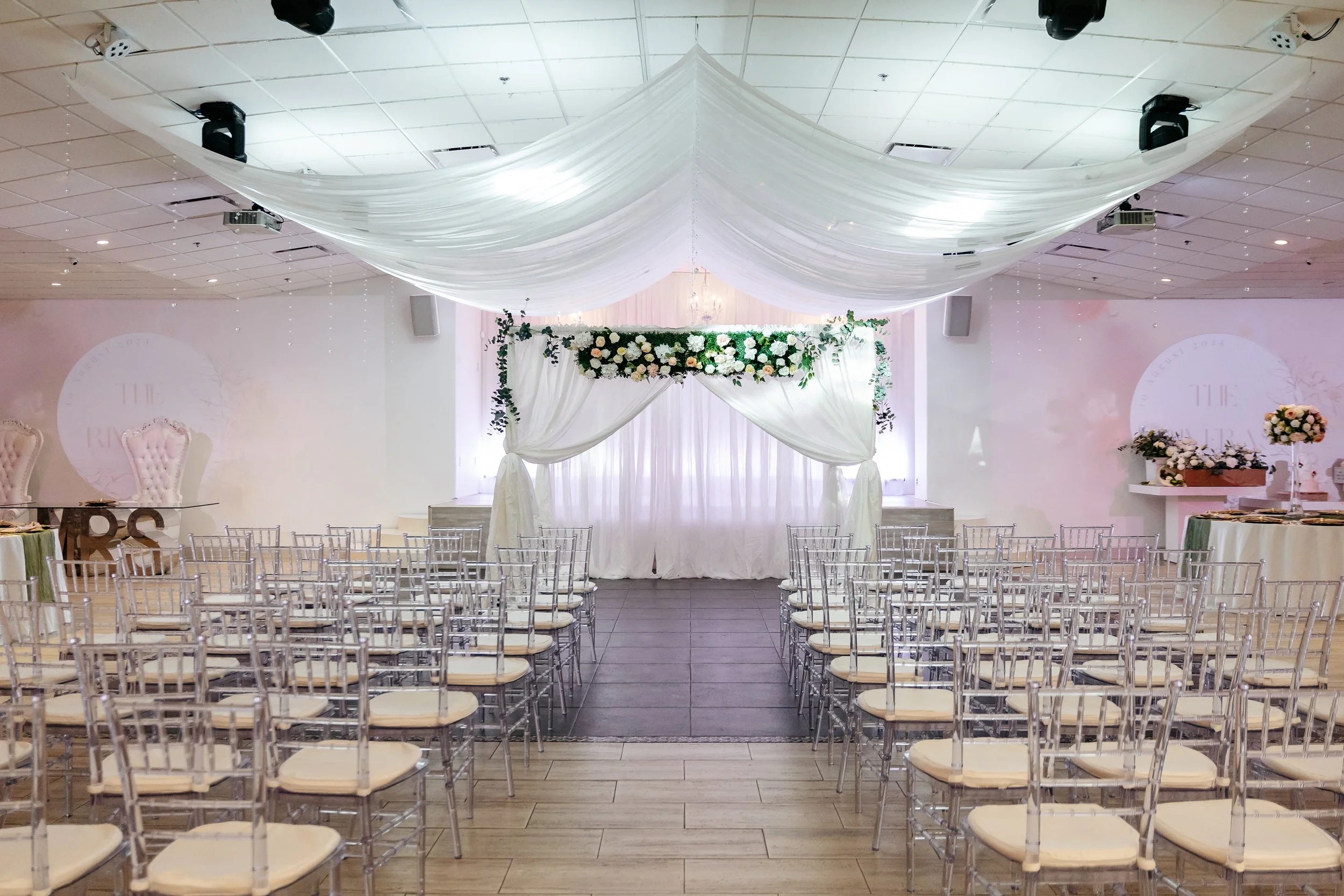 Wedding ceremony venue with rows of clear chairs, a decorated floral arch, draped fabric, and floral arrangements in a white and pastel color theme.