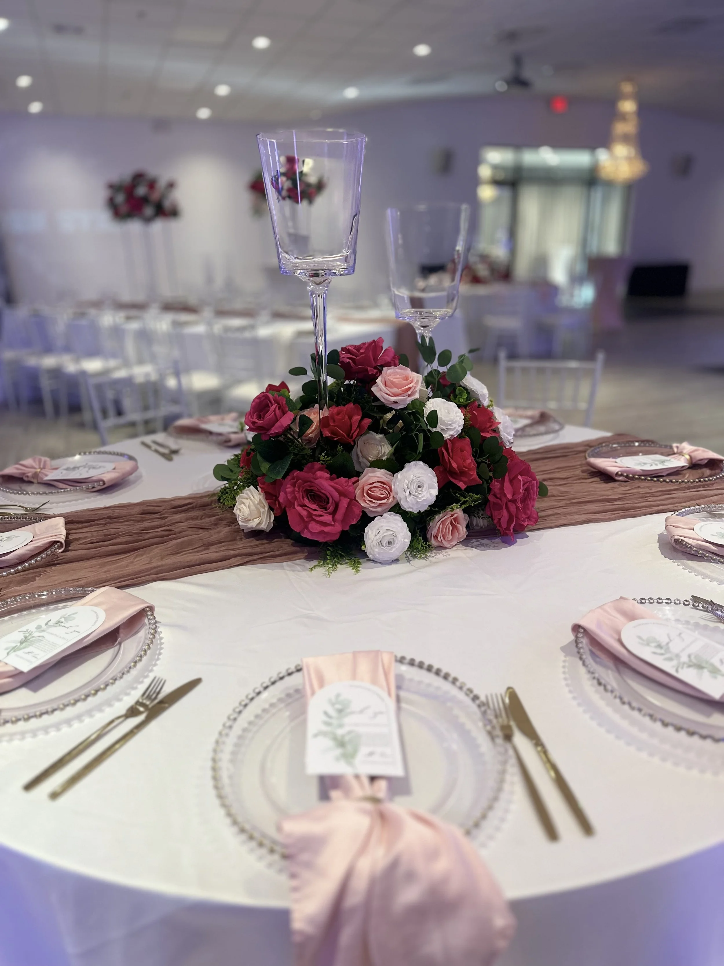 Elegant wedding table set with a floral centerpiece, pink and white roses, two tall glass candle holders, pink napkins with menus, gold cutlery, and white plates. Soft lighting and reception hall in the background.