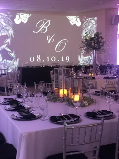 Elegant event reception with round tables, tall candle centerpieces, and a large floral arrangement. A projection on the wall displays initials 'B & O' and the date '08.10.19' in a decorative style.