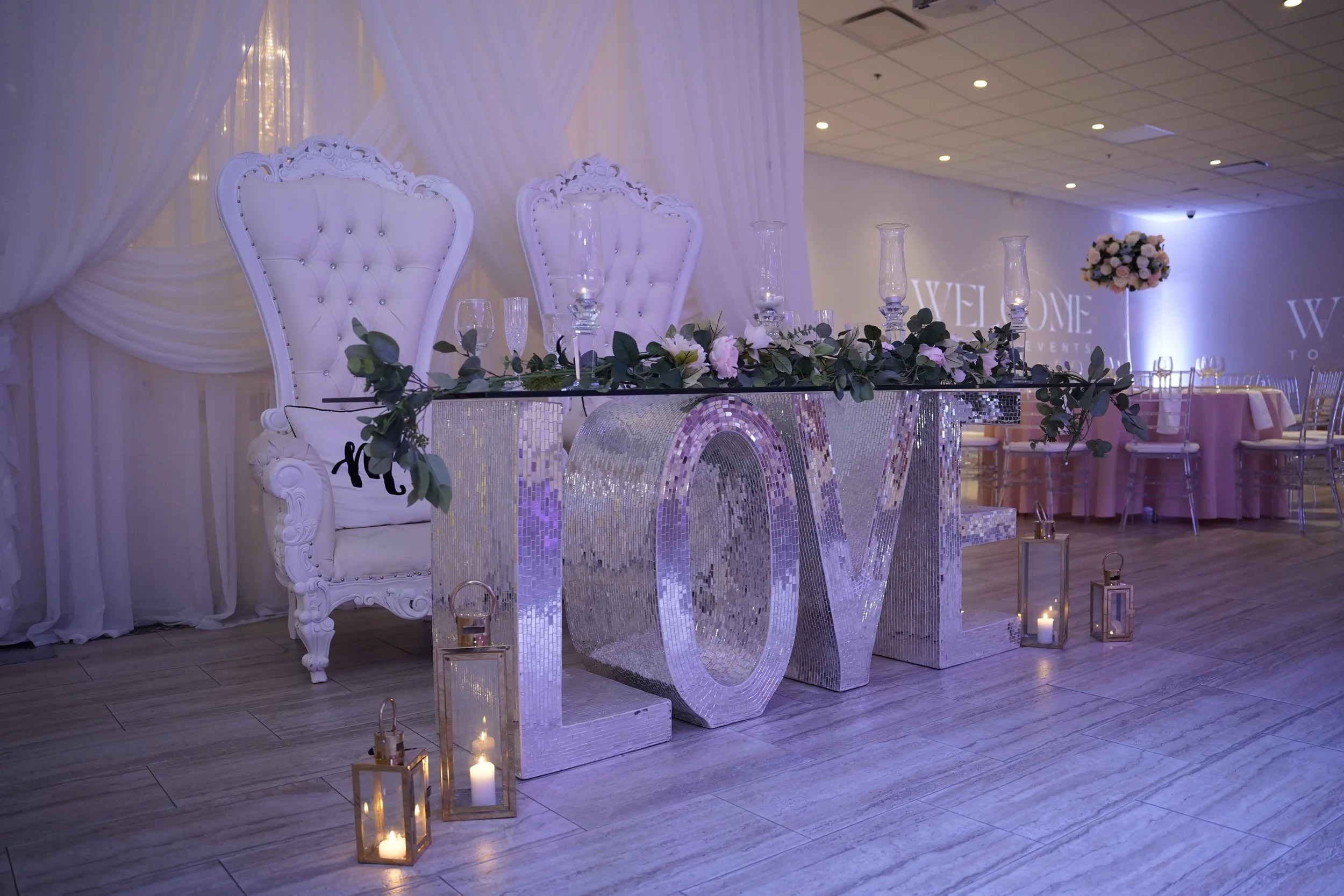 Elegant wedding reception table with large silver "LOVE" letters, floral garland, tall glass candle holders, candles in lanterns, and two ornate white chairs with tufted backs, set in a decorated hall with draped fabric and pink table settings in the background.