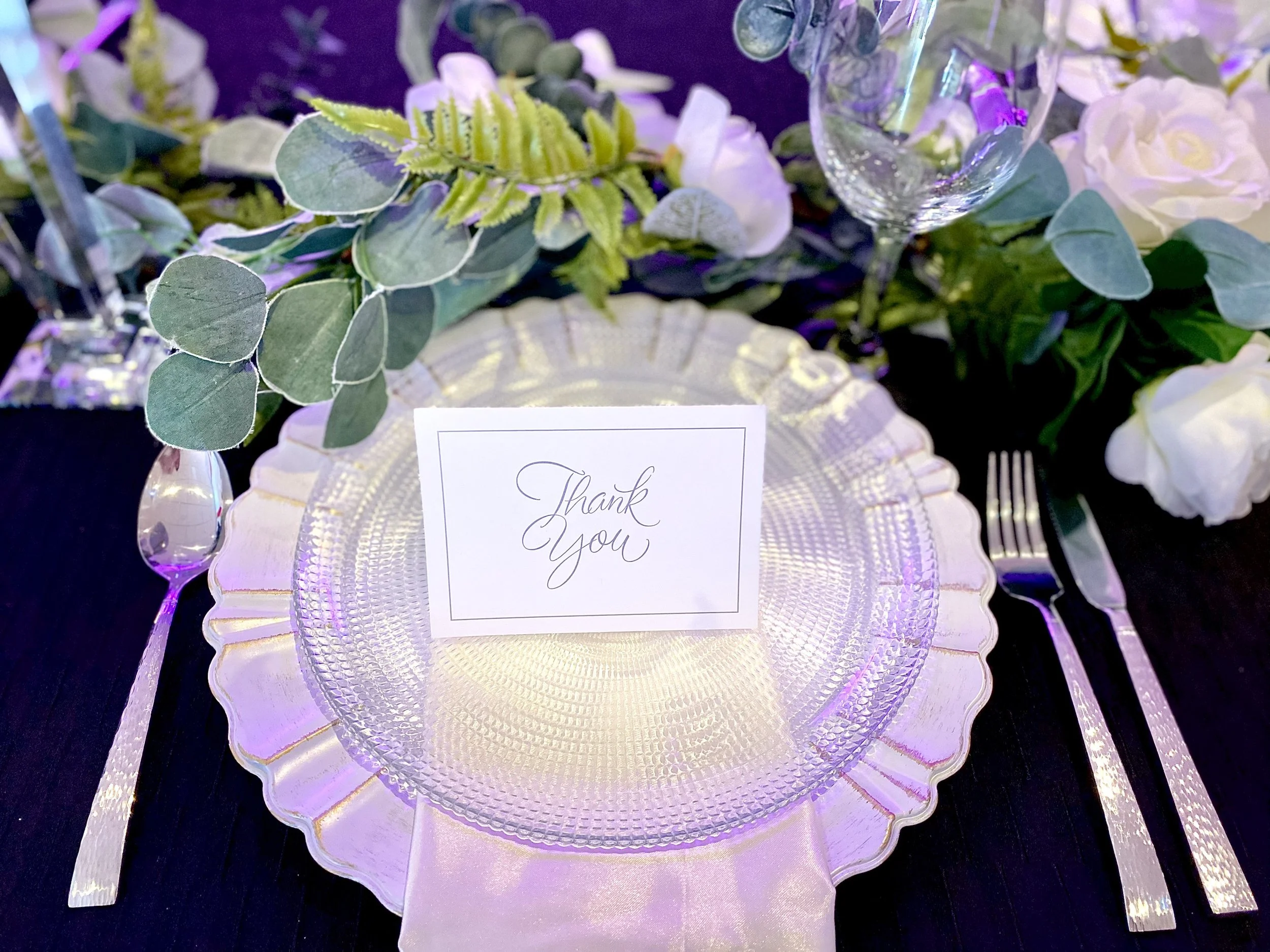 Elegant table setting with a clear textured glass plate, a white satin napkin underneath, a silver fork and knife on the right, a dessert fork on the left, a glass of water, and a