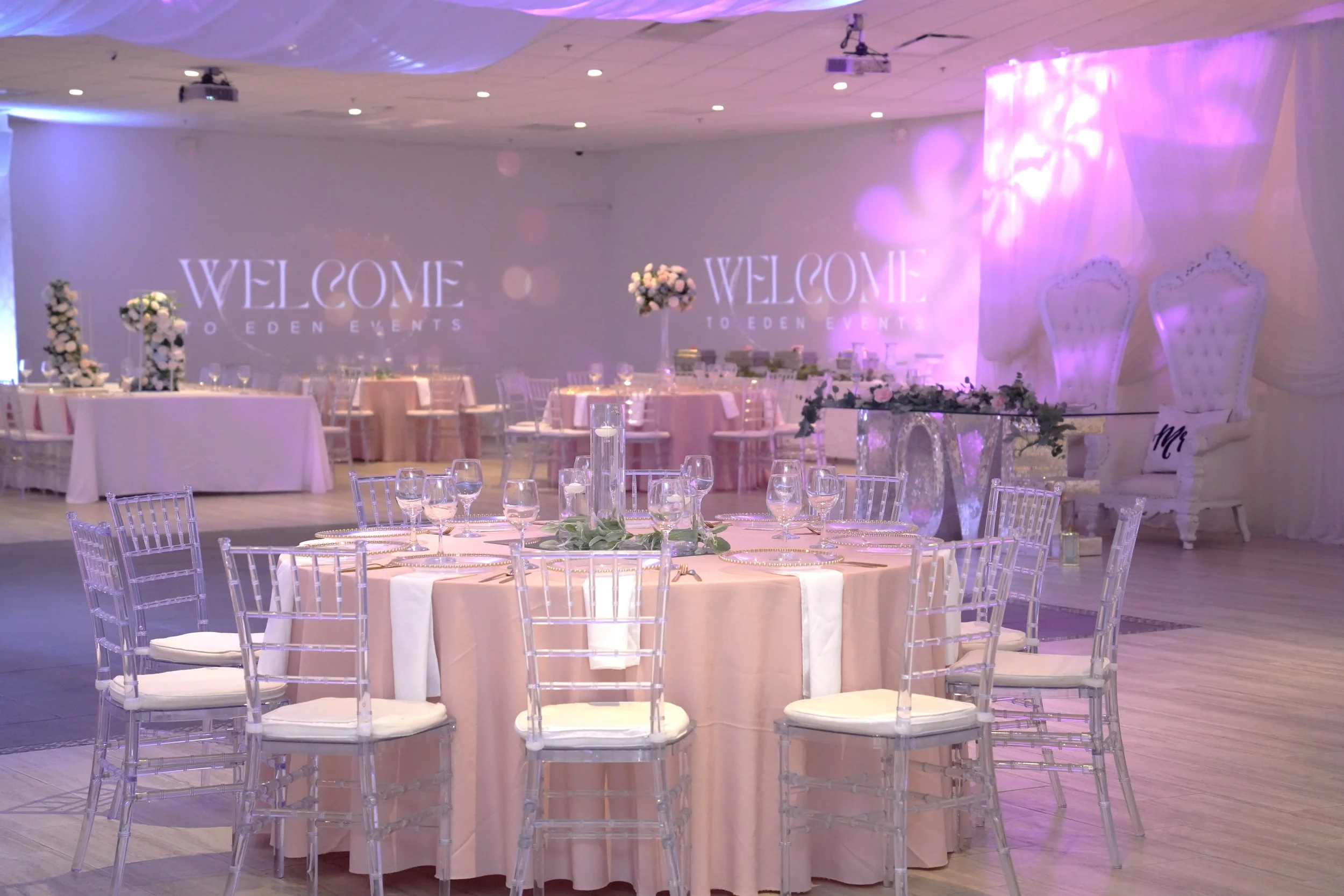 Elegant event decorated with pink and white flowers, round tables with pink tablecloths, clear chairs, with a backdrop displaying 'Welcome to Eden Events' in soft lighting, and two white ornate chairs at the side.