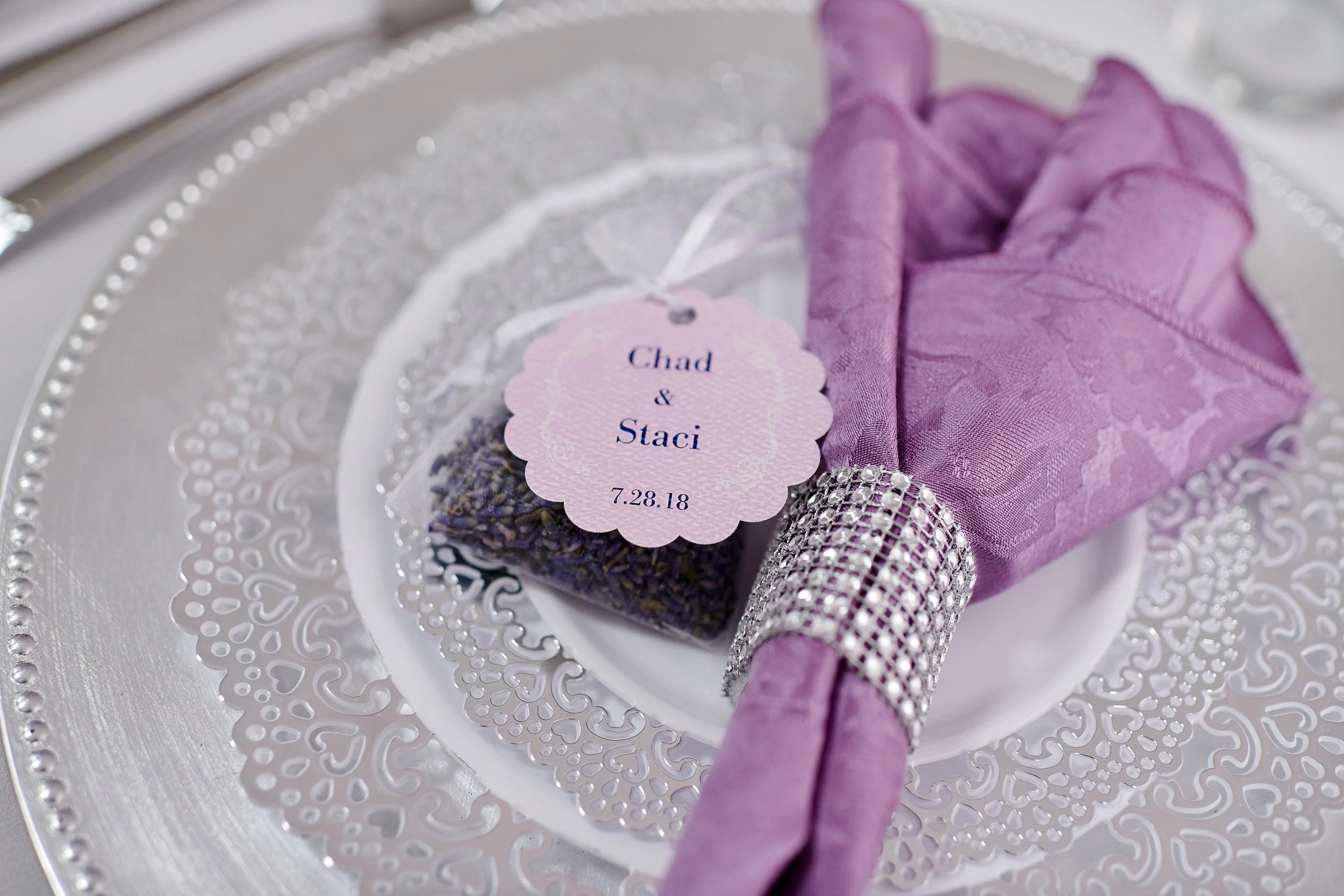 A decorative table setting featuring a silver ornate tray with a lavender lavender sachet wrapped in a purple cloth and adorned with a rhinestone band, a small bouquet of lavender, and a pink tag with wedding details for Chad and Staci, dated 7.28.18.