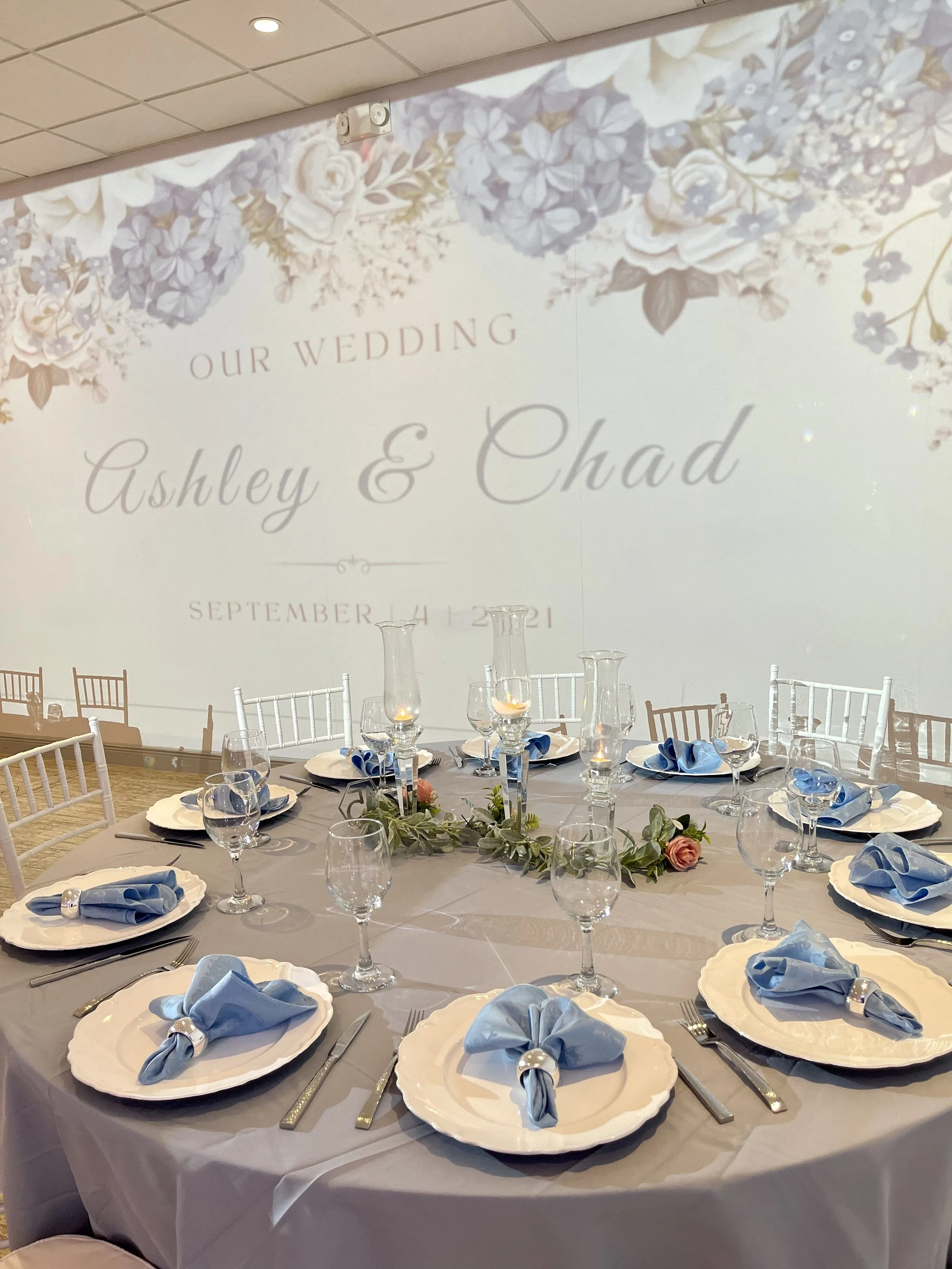 Wedding reception table with white plates, silverware, blue napkins, wine glasses, and a floral centerpiece. A large screen in the background displays the names Ashley and Chad and the date September 4, 2021.