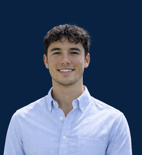 Young man with dark curly hair smiling, wearing a light blue button-up shirt, against a dark blue background.