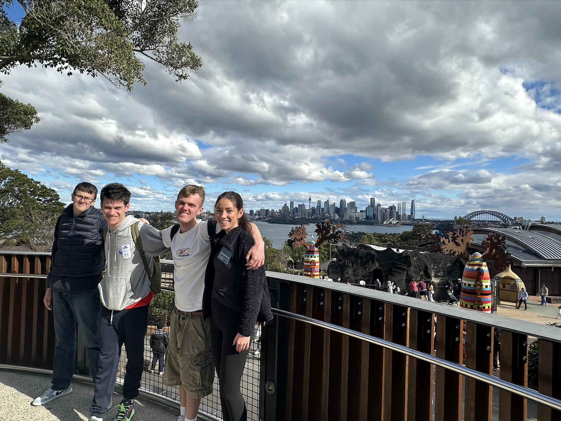 Four young adults standing together on a balcony overlooking a harbor in Sydney, Australia, with city skyscrapers, the Sydney Opera House, and Harbour Bridge in the background. They are smiling and posing for the photo, with some wearing backpacks.