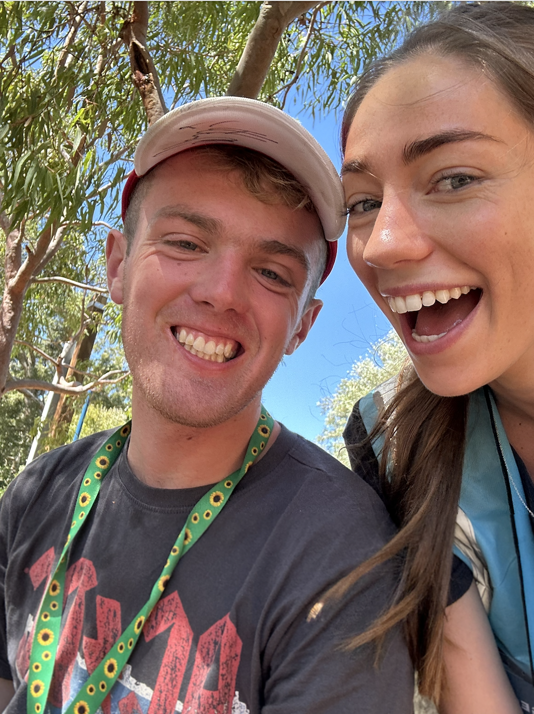 A close-up of a smiling young man and woman outdoors with trees and blue sky in the background.