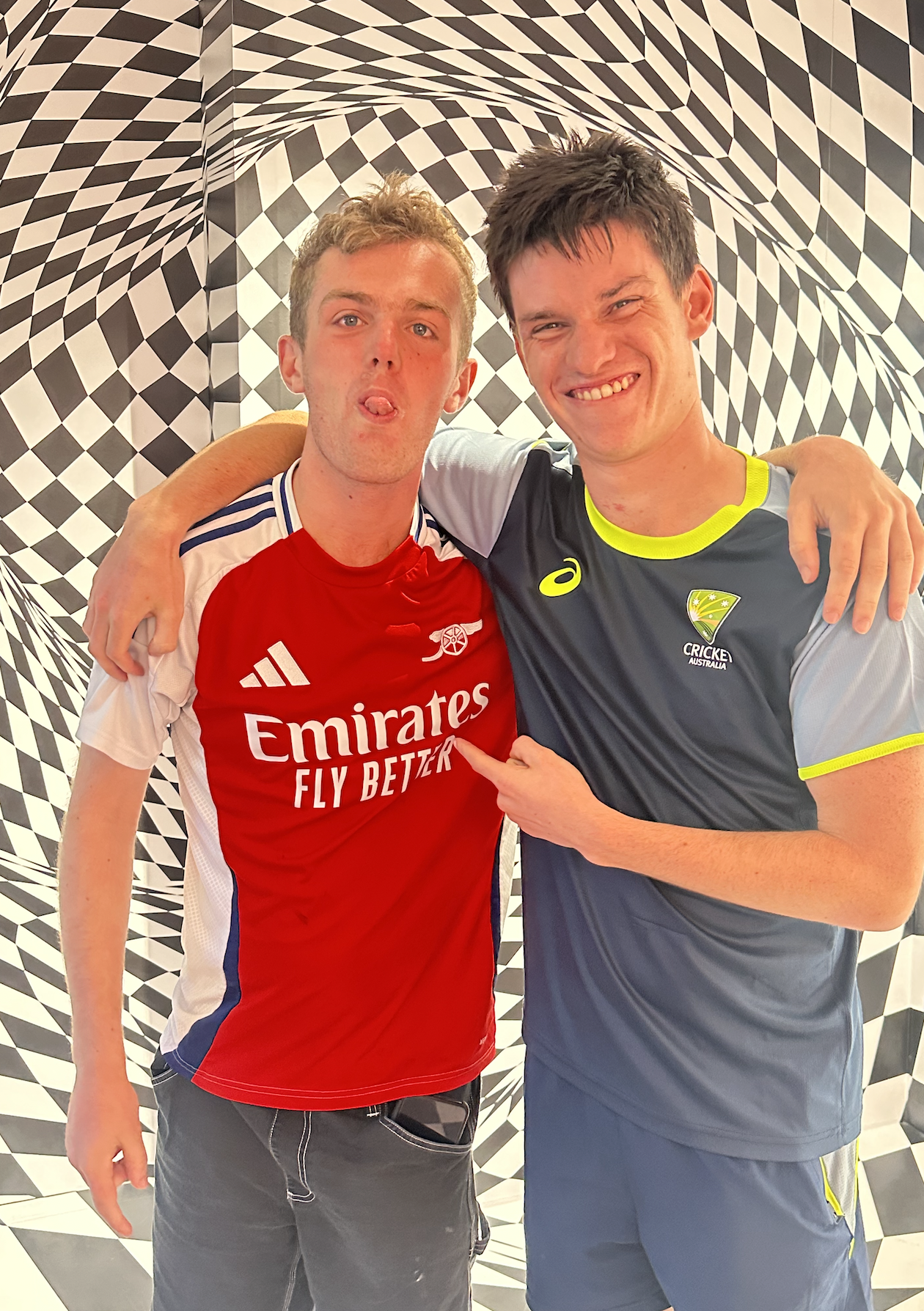 Two young men standing in front of a black and white optical illusion background, wearing sports jerseys from Arsenal and Cricket Australia, with one man pointing at the other's Arsenal logo.