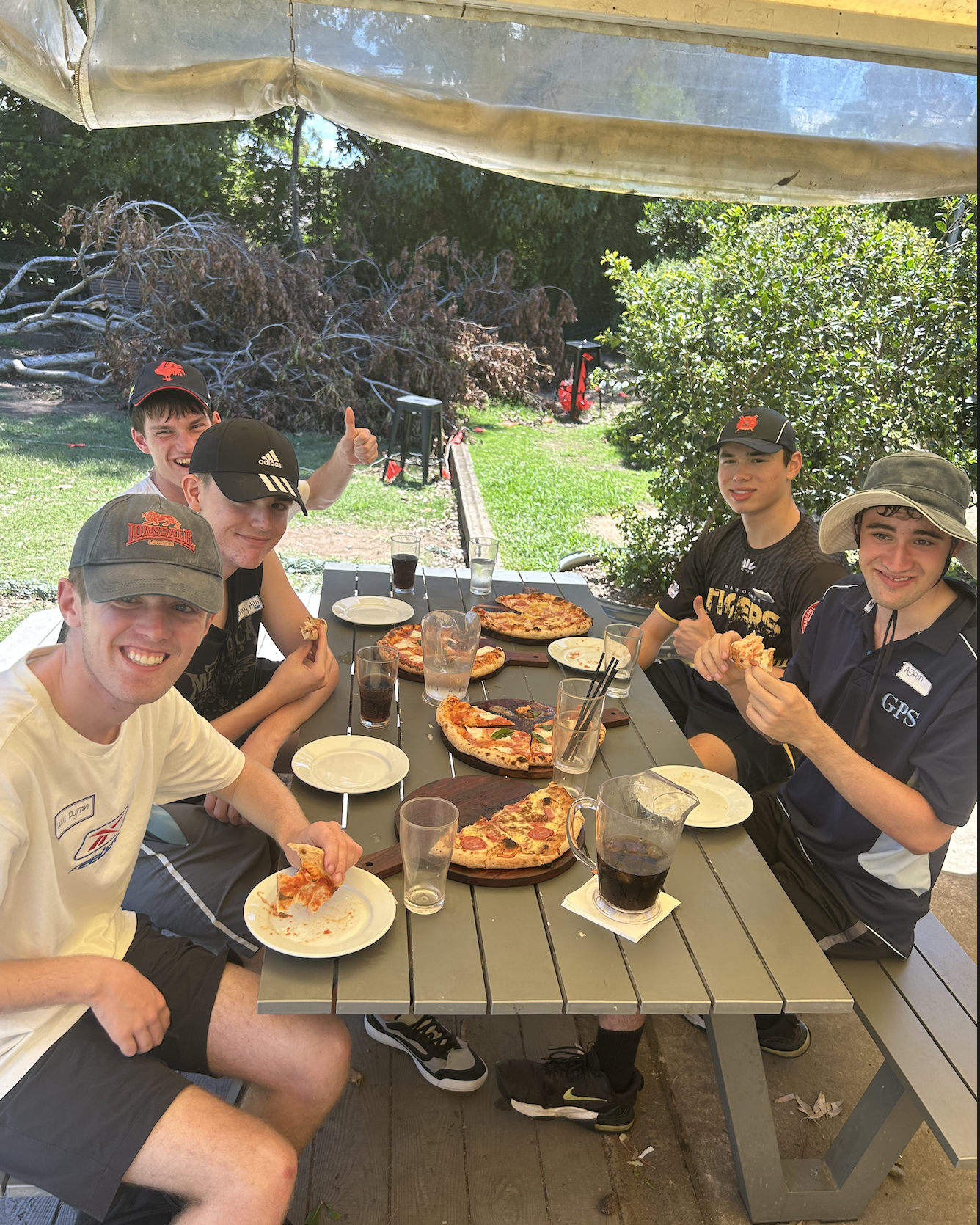 Five young men enjoying pizza and drinks at an outdoor dining table under a shaded canopy, with a garden and fallen tree branches in the background.