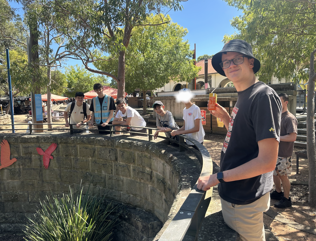 A group of young people standing around a circular stone pond with animated bird decorations, outdoors under trees on a sunny day, with a man in a black hat and glasses holding an orange drink in the foreground.
