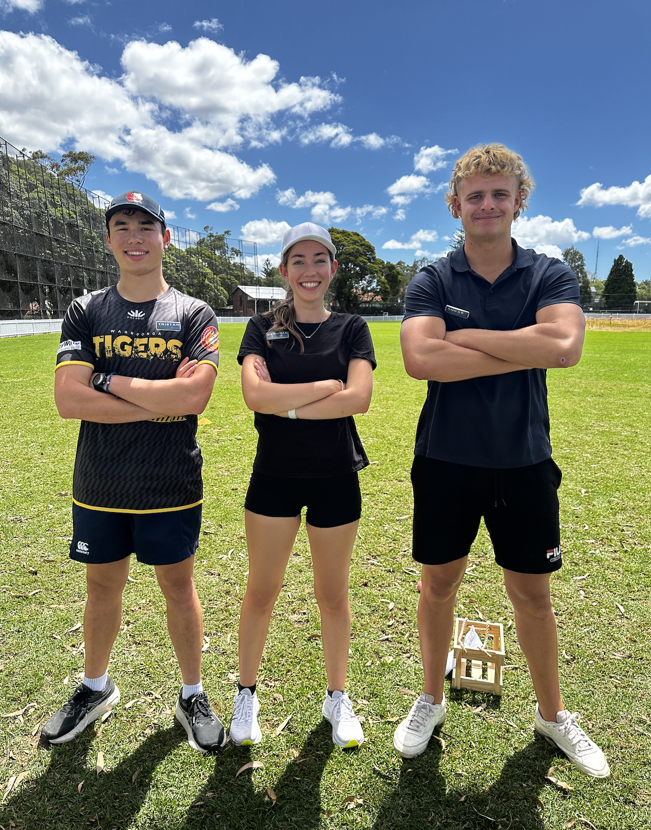 Three young athletes standing on a grassy field with arms crossed, under a blue sky with clouds. The person on the left wears a black sports jersey and shorts, the person in the middle wears a black t-shirt and shorts, and the person on the right wears a black sports shirt and shorts. There is a wooden box on the ground near the right athlete.