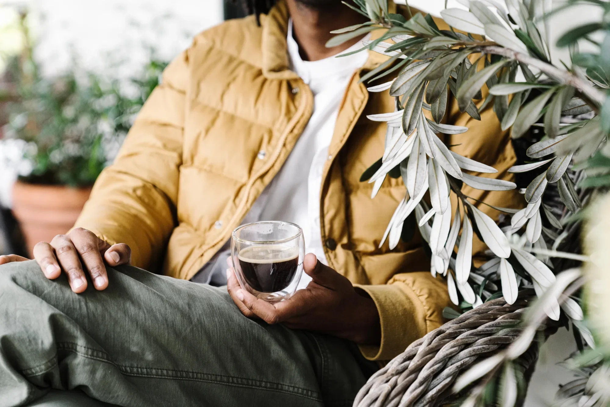 Person in a mustard puffer jacket holding a glass cup of coffee beside an olive branch