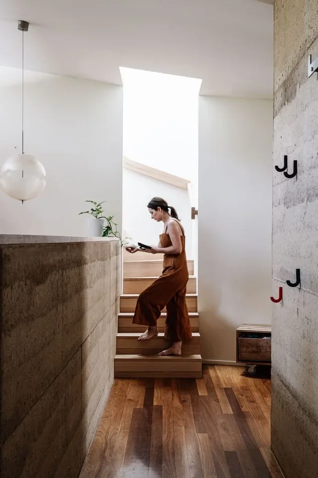 Woman reading on a timber staircase beside a rammed-earth wall, lit from a skylight above.
