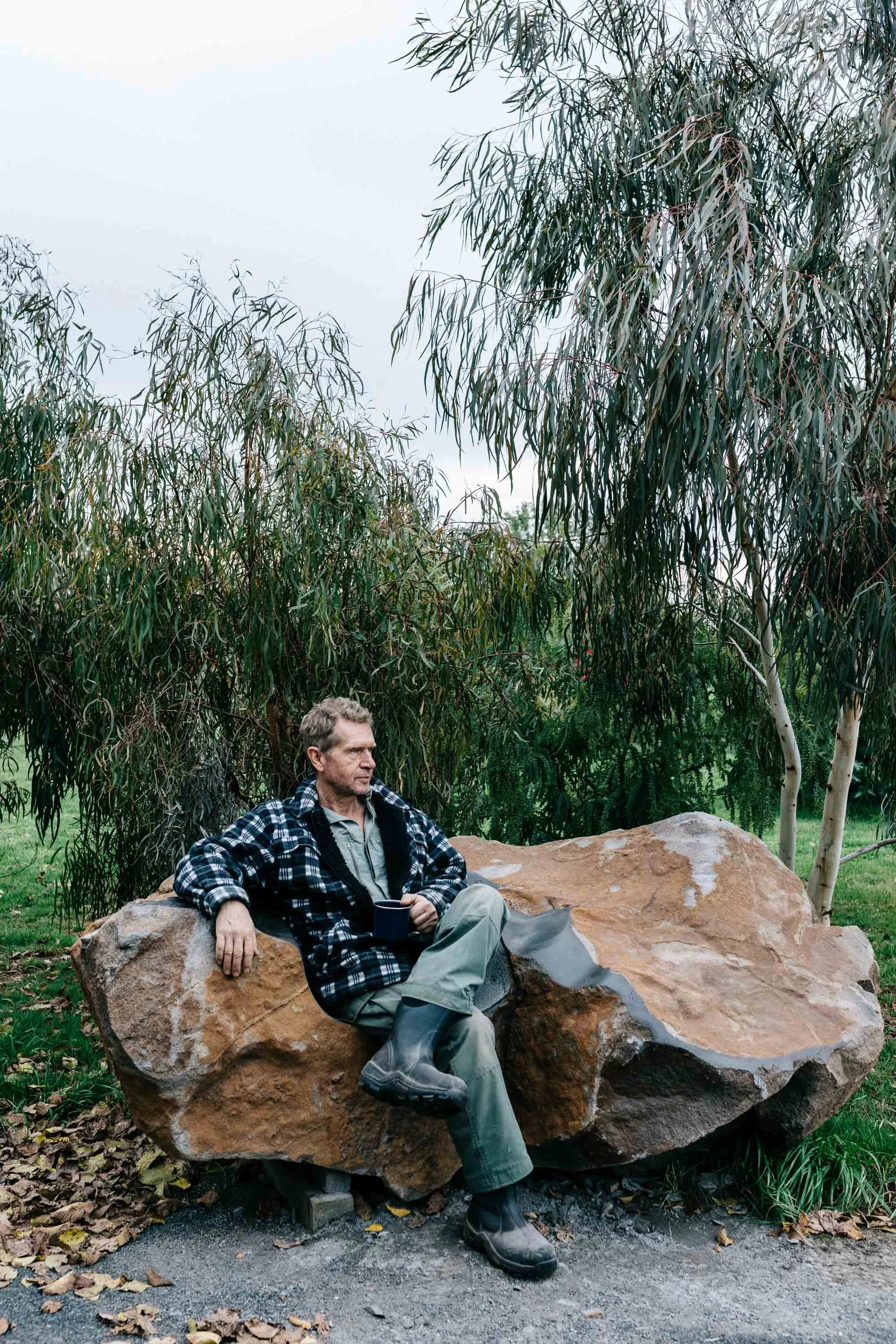 Man in plaid shirt with a coffee sitting on a giant rock that doubles as habitat