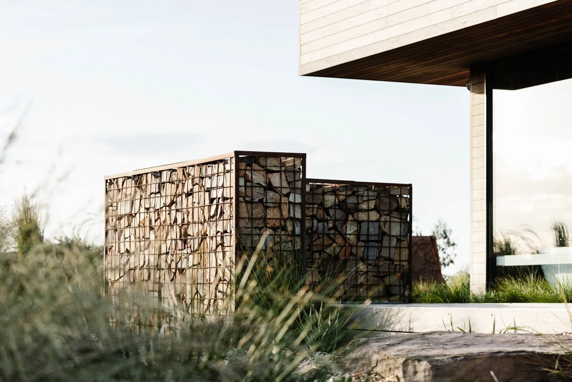 Rock-filled gabion cages in grasses beside the timber-lined corner of Elemental House.