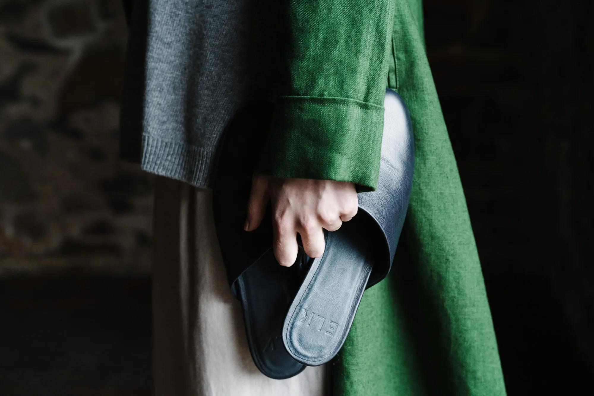 Person in a green ELK the Label coat holding a pair of black slippers