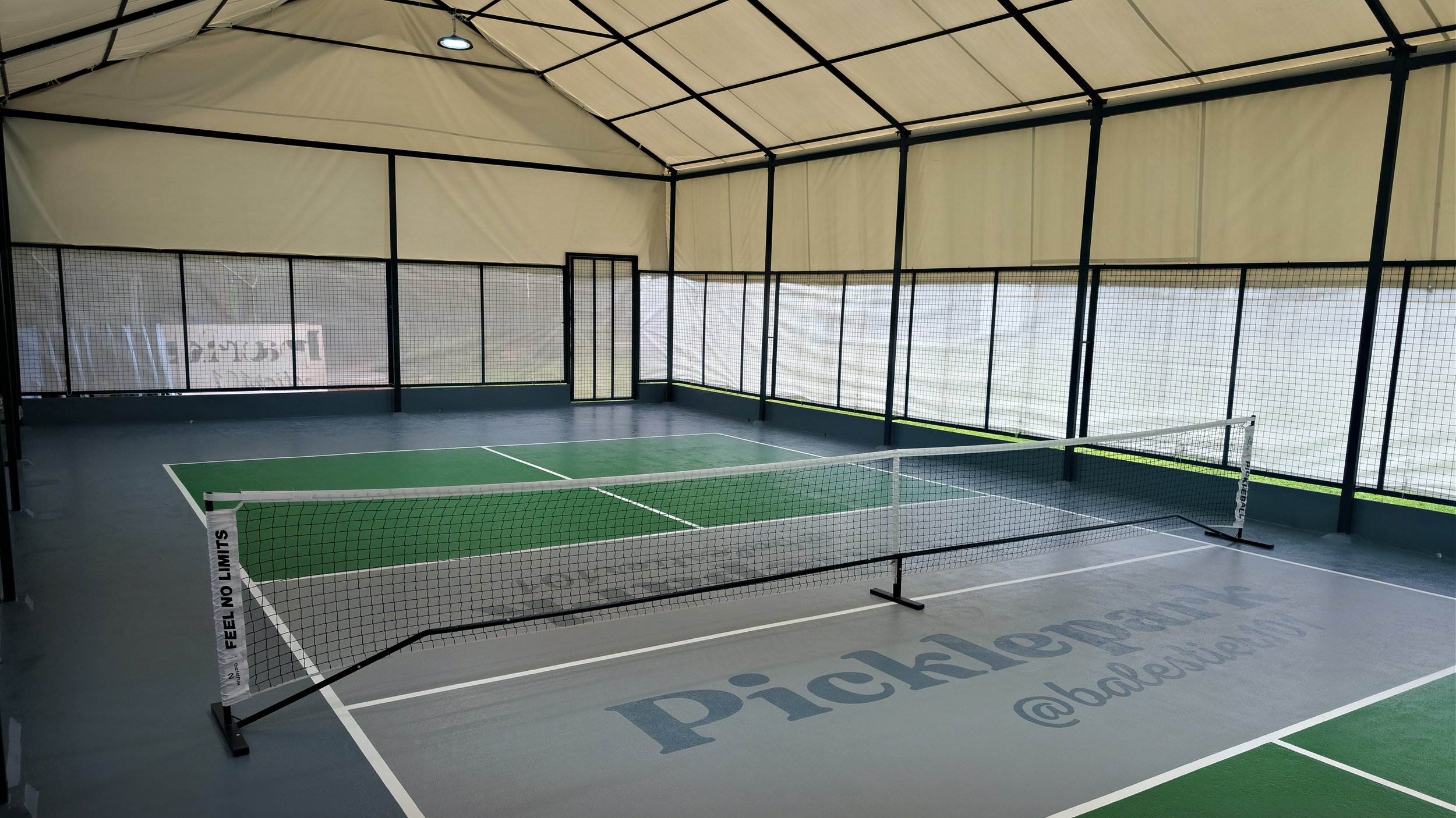 Indoor pickleball court with green and gray surface, net setup, and protective netting around the perimeter.