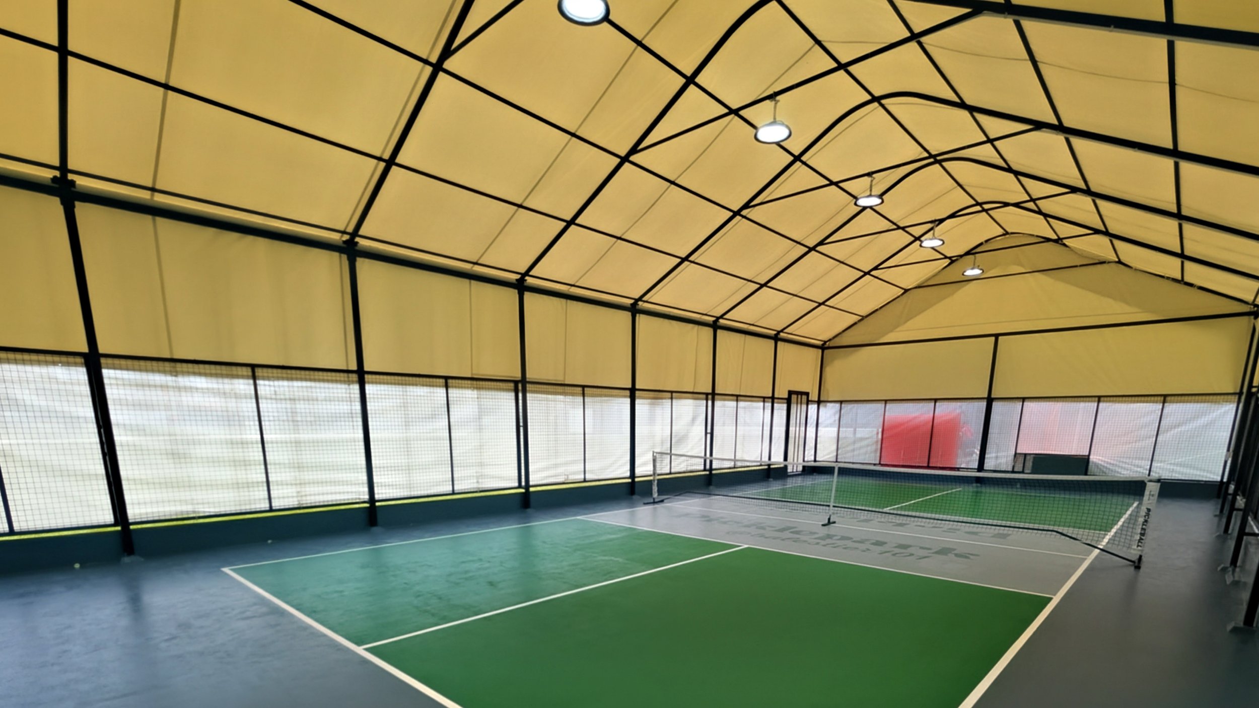 Indoor tennis court with green and blue flooring, surrounded by a tall net and enclosed with a yellow ceiling and walls, illuminated by overhead lights.